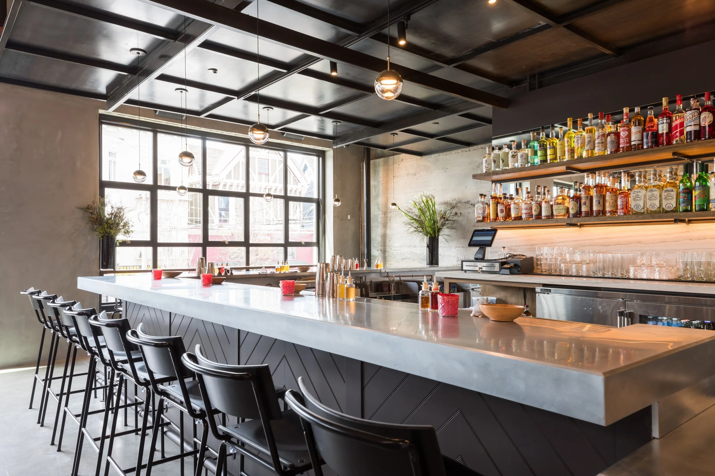 Che Fico restaurant in San Francisco, California. Modern bar with a row of black chairs at a white marble countertop, bottles of alcohol on wooden shelves, large windows allowing natural light, and ceiling lights with glass globes.