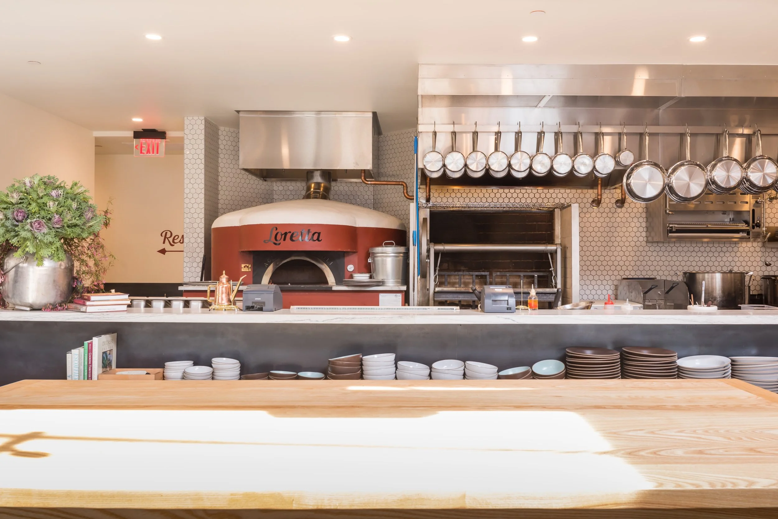 Che Fico restaurant in San Francisco, California. Kitchen area with hanging pots and a wood-fired pizza oven labeled 'Loretta'. A counter with various dishes, books, and a large flower arrangement to the side.