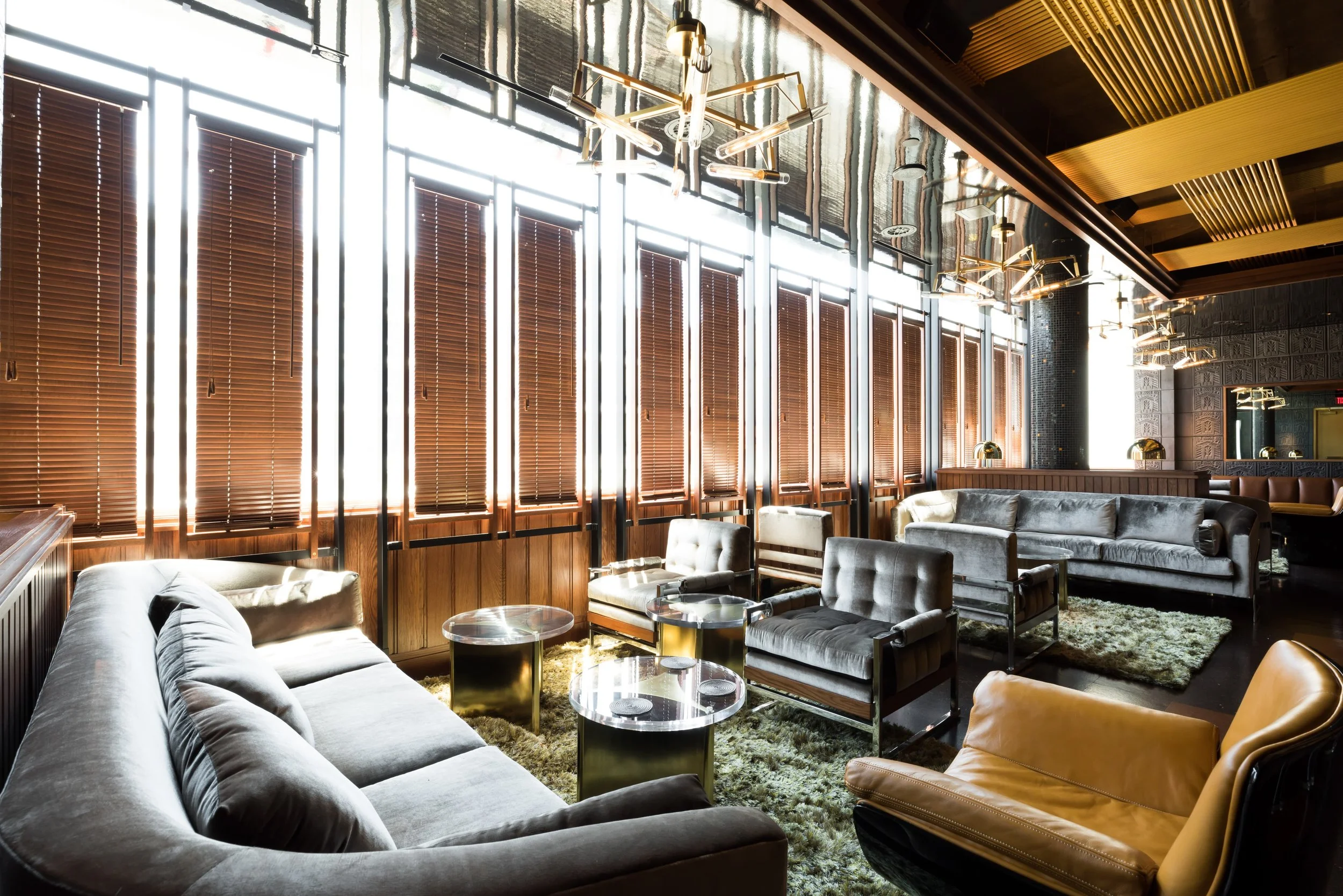 A modern lounge with large windows covered by wooden blinds, stylish seating including a sofa and armchairs, glass coffee tables, a plush rug, and artistic light fixtures on the ceiling.