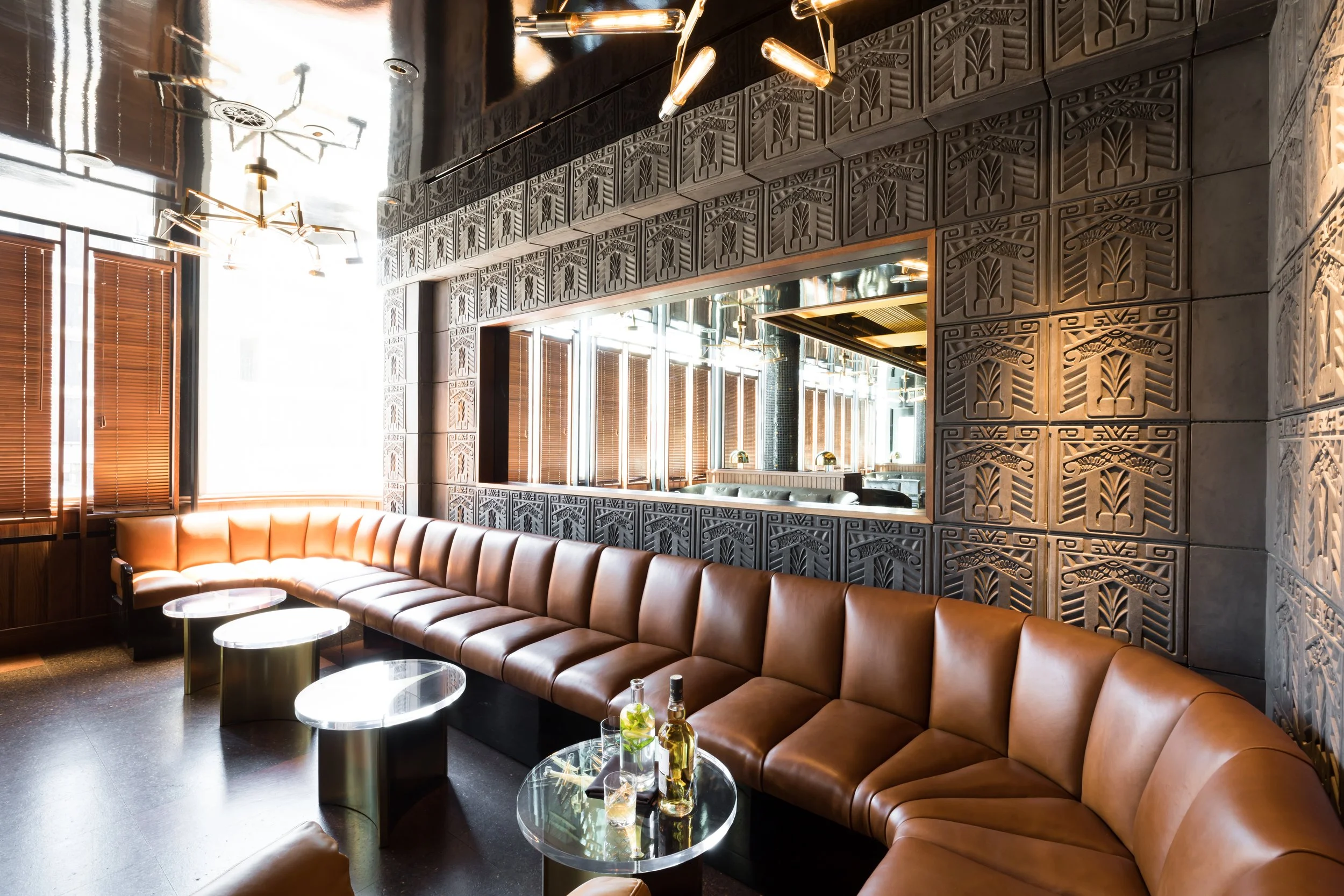 Cold Drinks bar above China Live Market in San Francisco, California. Modern interior with a curved brown leather sofa, small round glass coffee tables, decorative dark textured wall, large mirror, and stylish ceiling light fixtures.