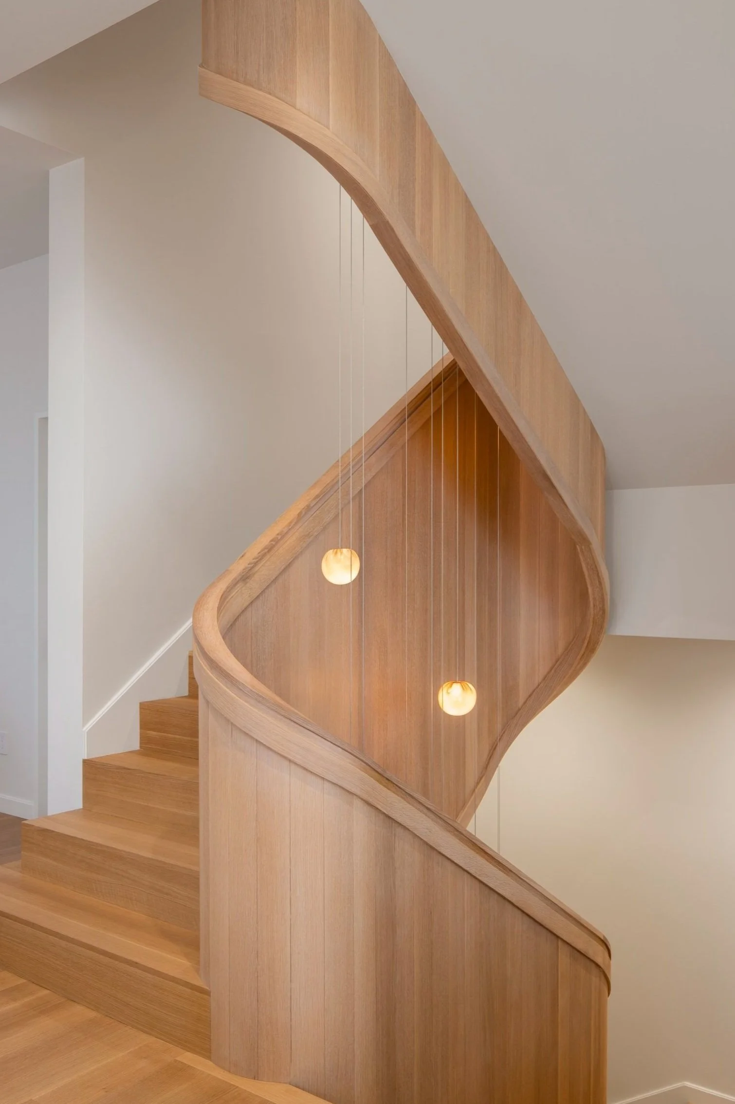 Six-Story City House in San Francisco, California. A wooden staircase with a curved handrail and vertical wooden slats. Two spherical pendant lights hang next to the staircase, illuminating the space.