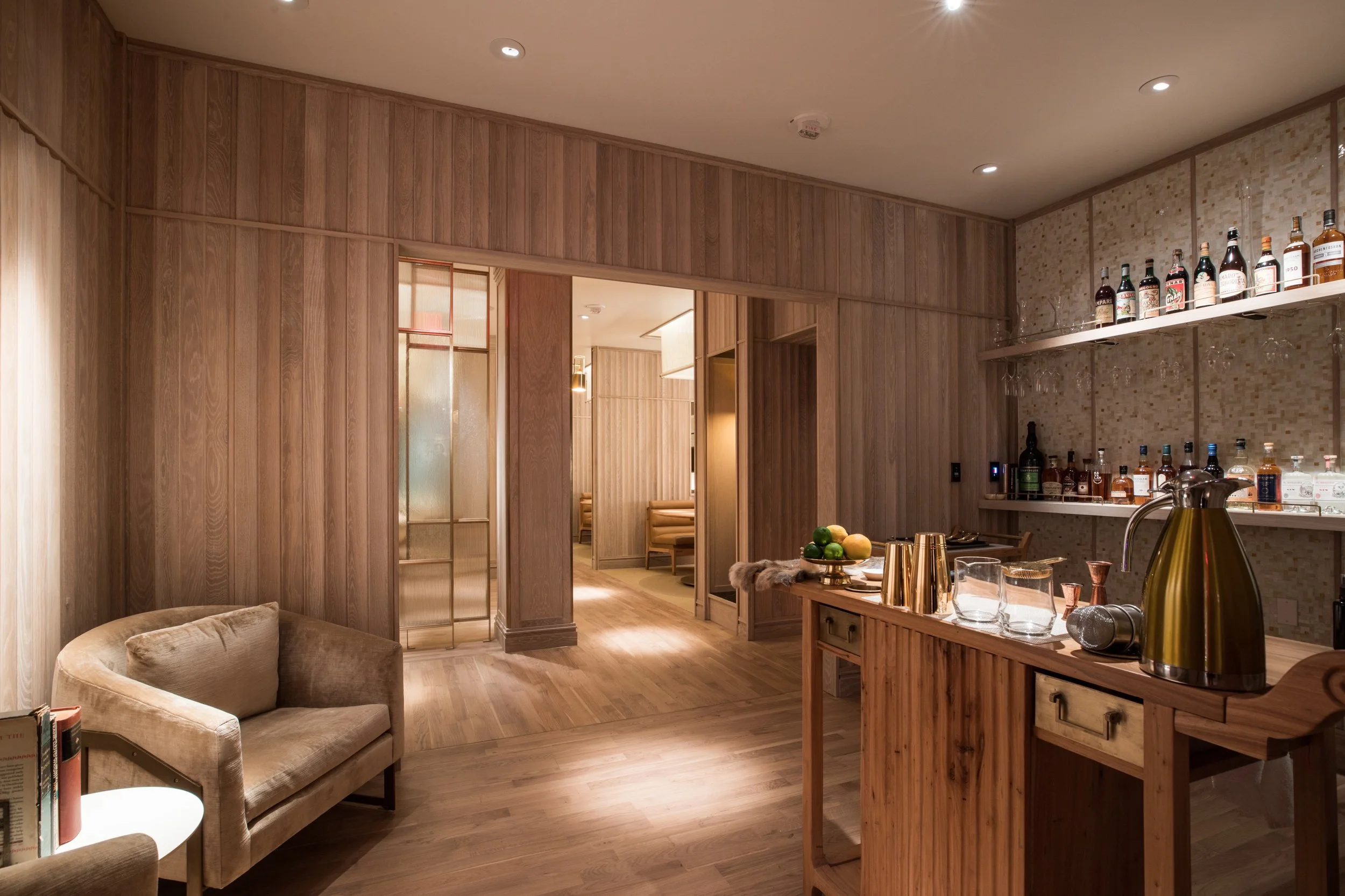Eight Tables restaurant above China Live Market in San Francisco, California. Interior of a cozy room with wood-paneled walls, a beige sofa, a side table with books, and a bar area with bottles of alcohol and glassware, illuminated by warm lighting.