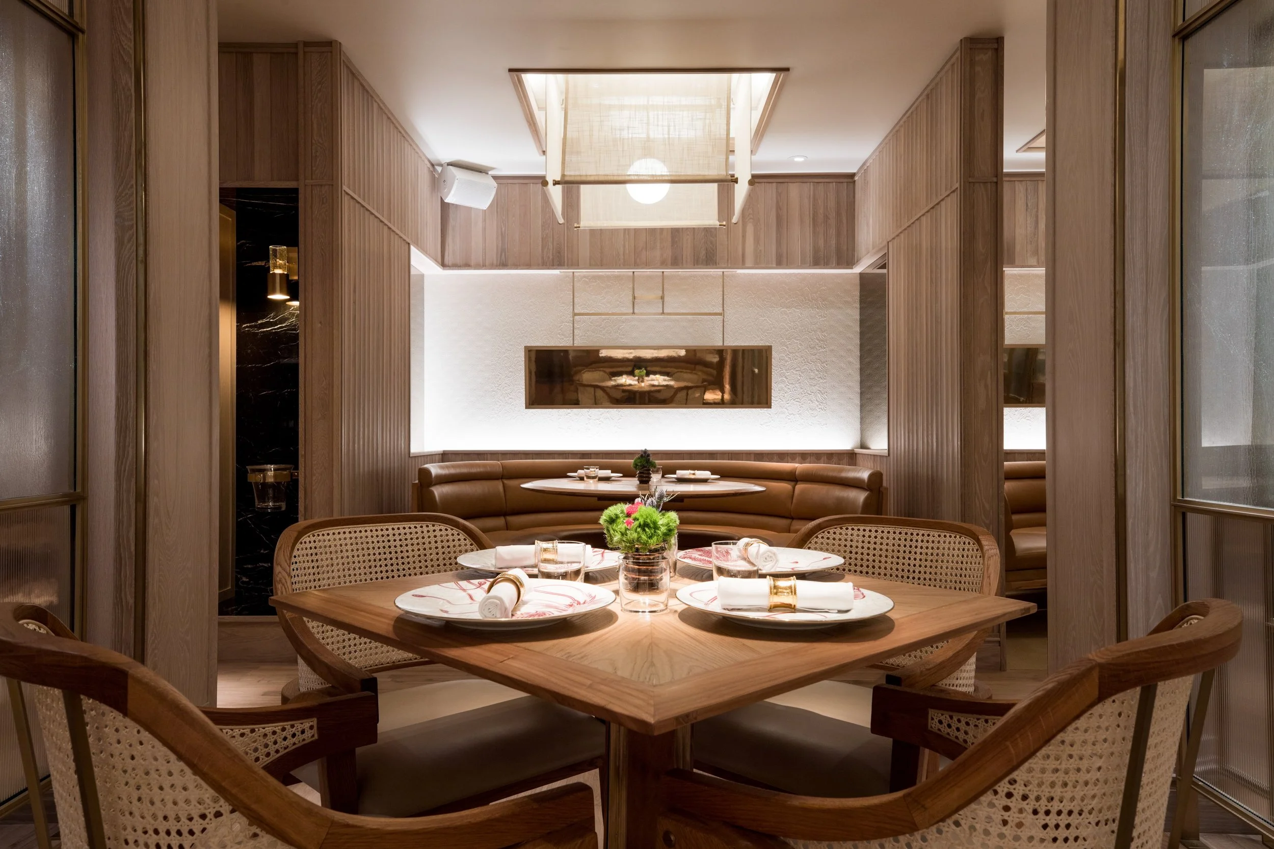 Eight Tables restaurant above China Live Market in San Francisco, California. Dining room with a round table set for four, beige woven chairs, a small centerpiece, and a wooden wall with a large mirror and a hanging light fixture overhead.