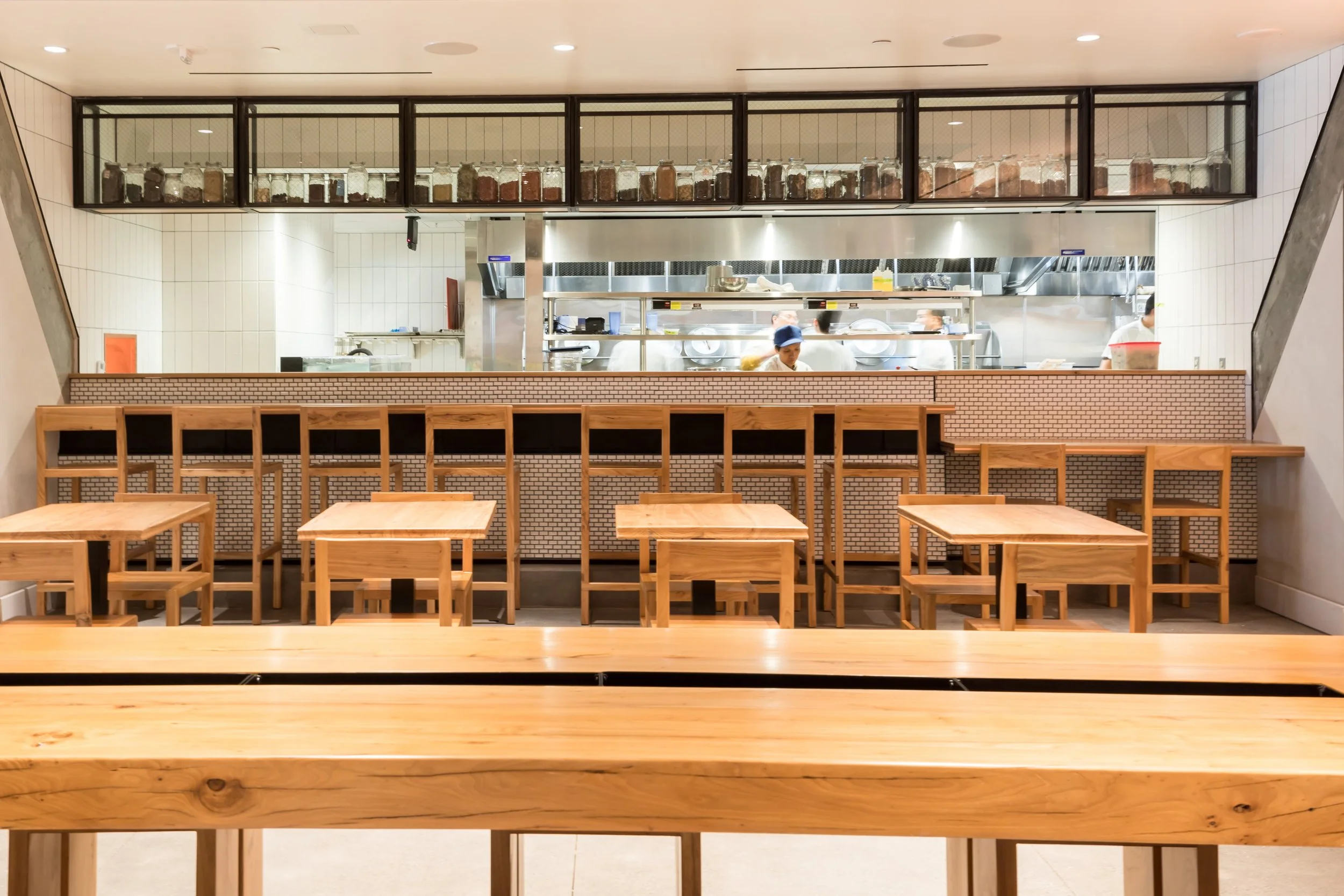 China Live Market in San Francisco, California. Wooden tables and chairs in a restaurant with a view of the kitchen behind a counter. The kitchen has chefs and jars on a shelf above.