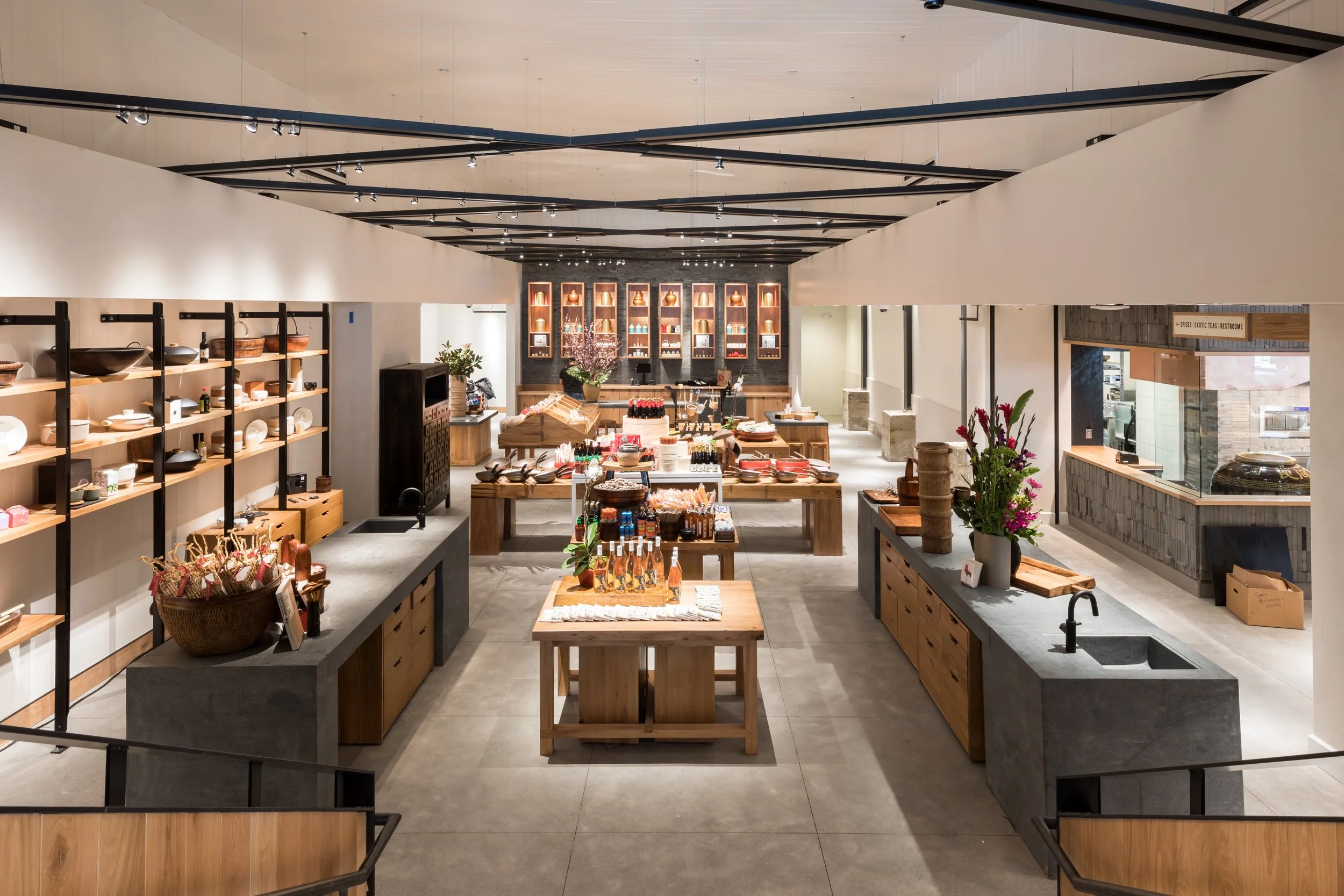 China Live Market in San Francisco, California. Experiential Retail. Interior of a store with wooden shelves and tables displaying various products, decorated with potted plants and flowers, with black ceiling beams and track lighting.
