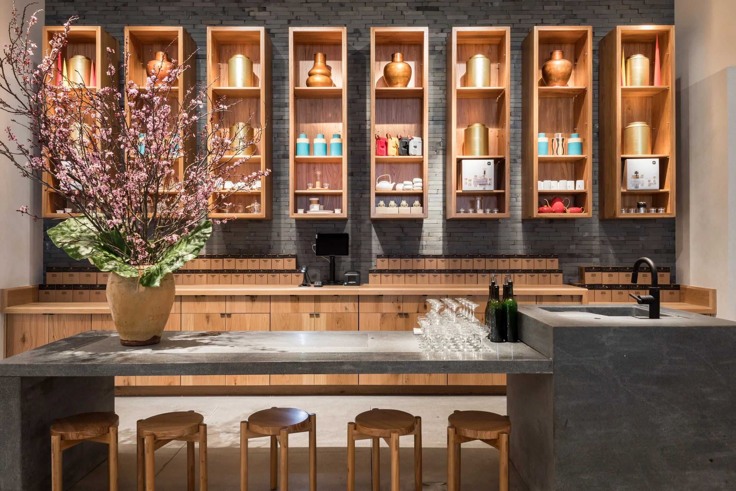 A modern kitchen with black brick wall, open wooden shelves with vases and containers, a potted pink flowering plant on a gray marble counter, bottles, glasses, and a sink.