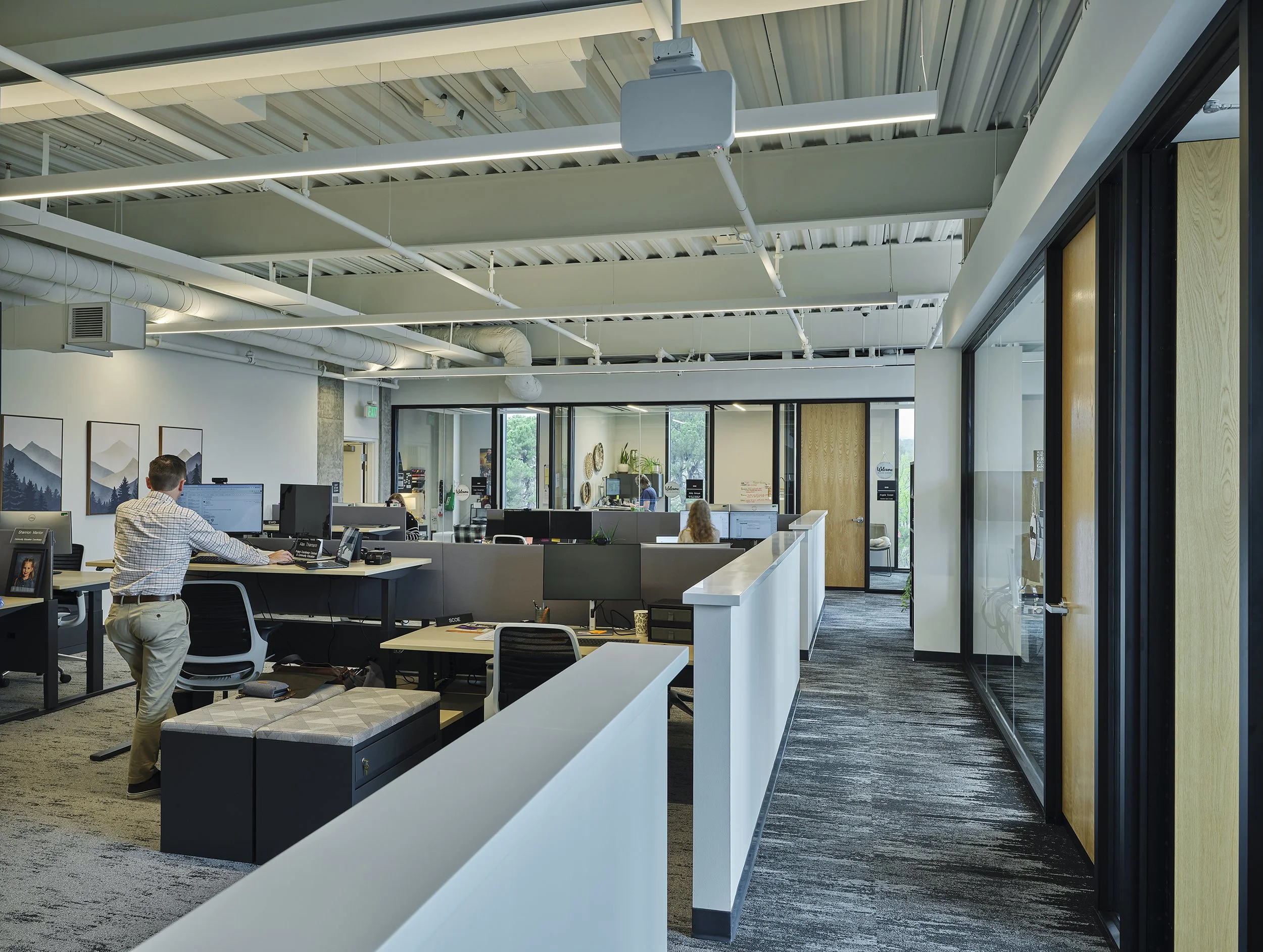 Shasta College in Redding, California. Part of Block 7 Development. Open-plan modern office with employees working at desks, large windows, exposed ceiling with industrial piping, and artwork on the walls.