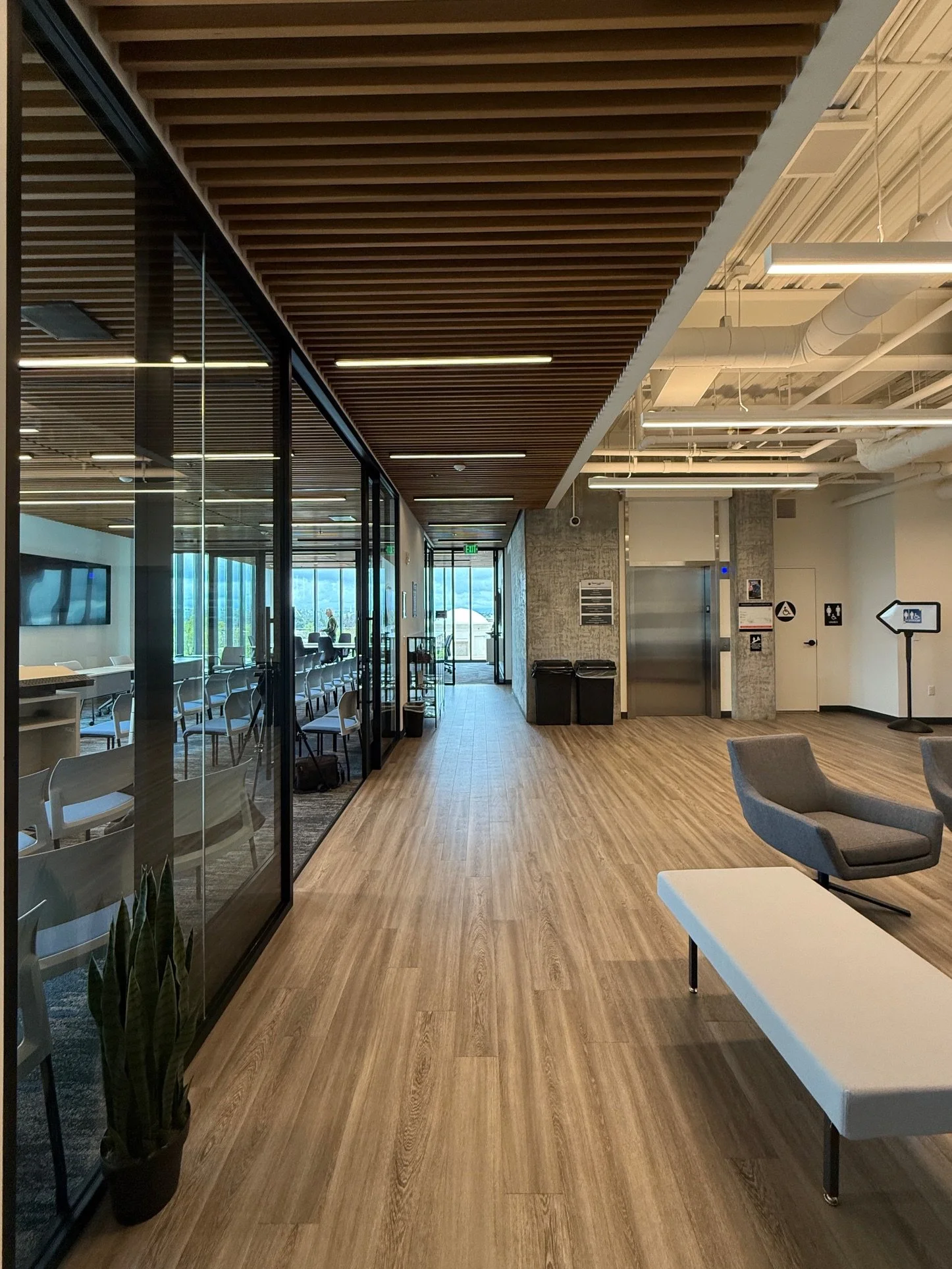 Shasta College in Redding, California. Part of Block 7 Development. Empty modern waiting area or lounge with wooden floors, a white table, gray chairs, glass walls, and a view of the outside with cloudy sky and trees.