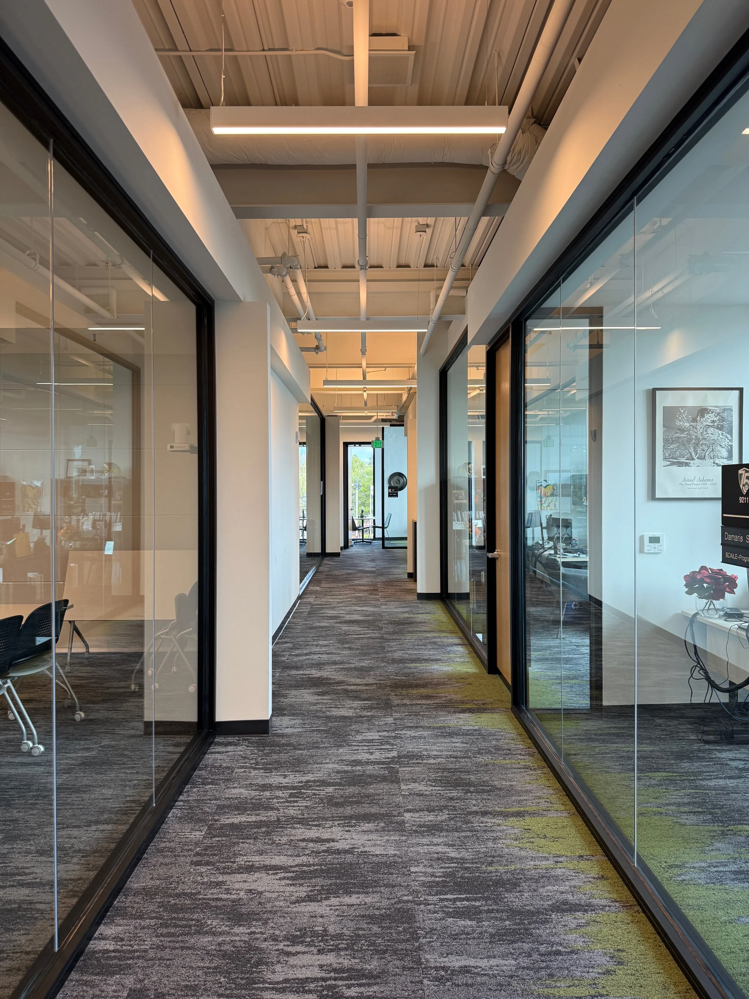 Shasta College in Redding, California. Part of Block 7 Development. A hallway inside an office building with glass walls on both sides, hardwood flooring, and a ceiling with exposed piping and modern light fixtures.