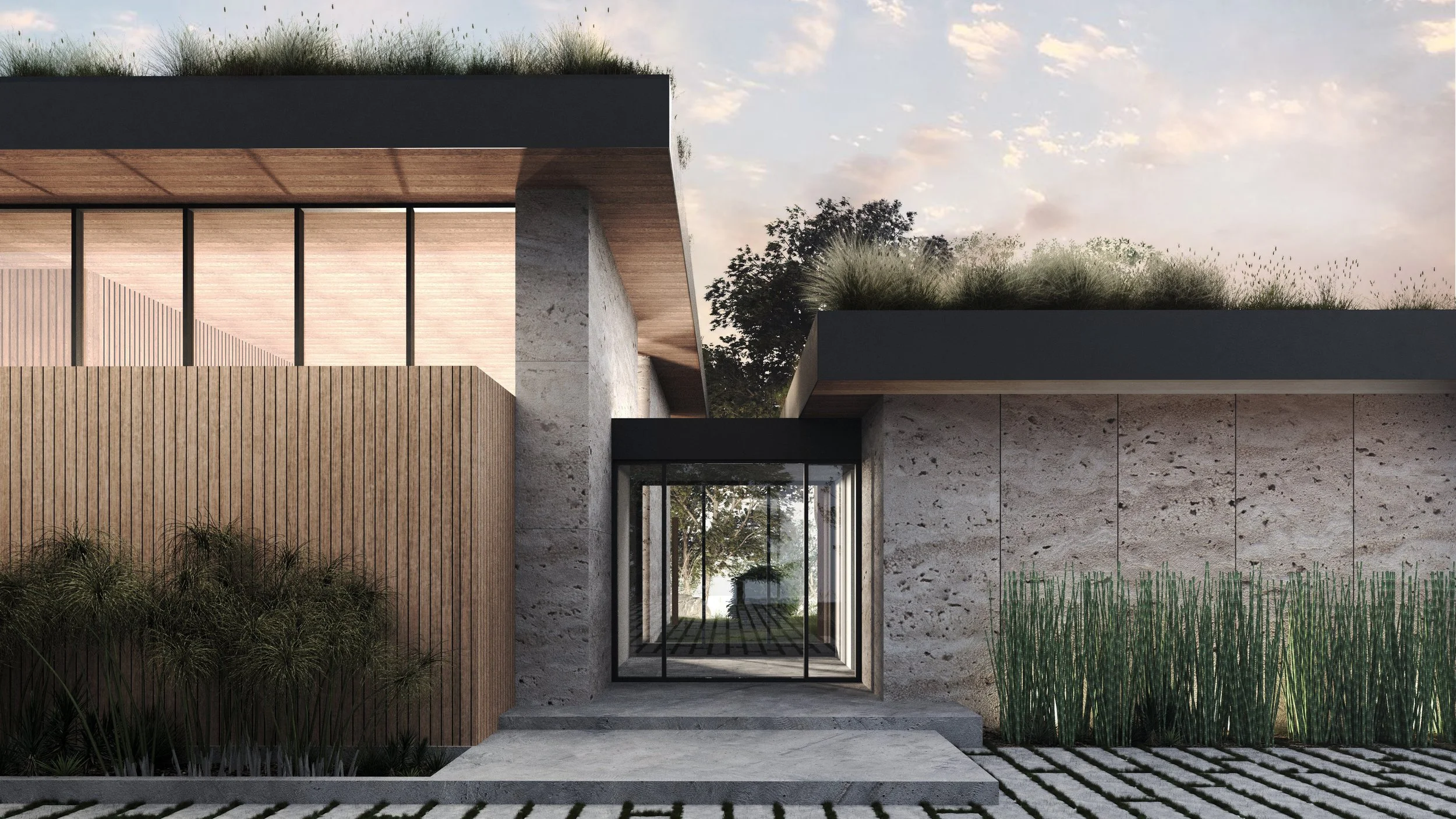 Carmel Valley Hillside Home in Carmel, California. Modern building exterior with stone, wood, and black accents, large glass doorway, and landscaping including tall grasses and trees in the background, under a partly cloudy sky.