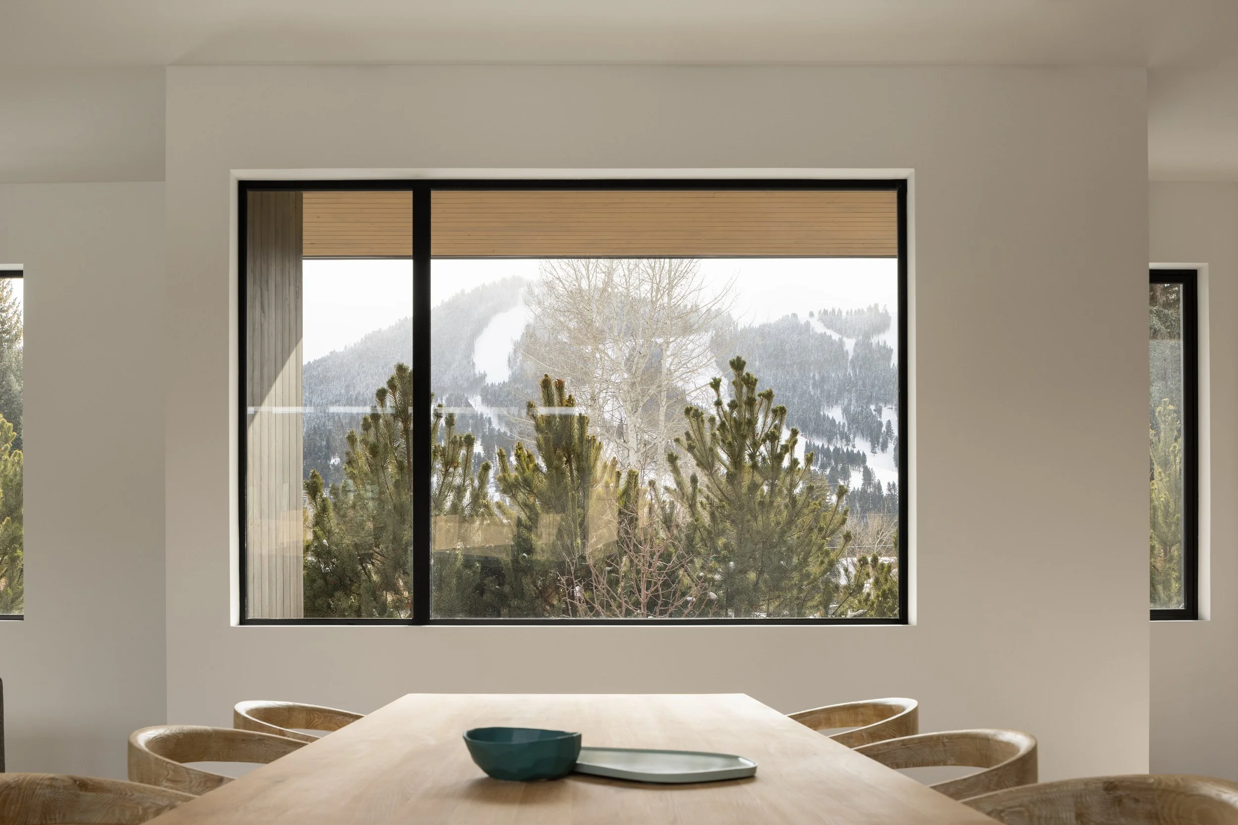 A modern dining room with a large window showing a snowy mountain landscape and trees outside. The room has light-colored walls, a wooden dining table, and wooden chairs. A bowl and tray are on the table.