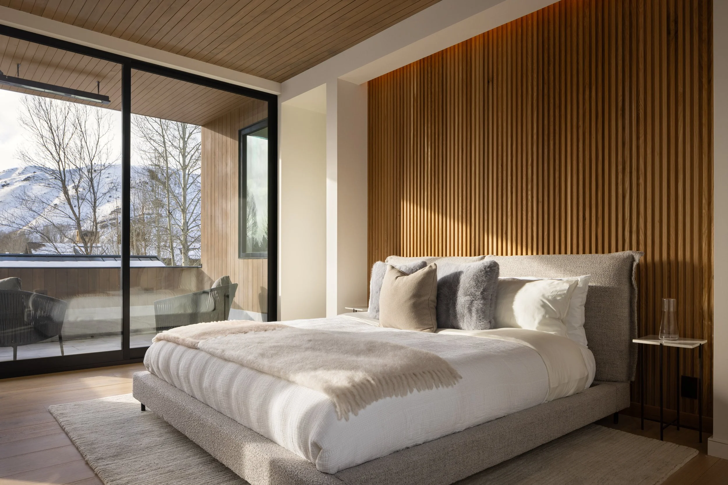 Teton House in Jackson, Wyoming. Modern bedroom with large bed, neutral-colored pillows, and cozy blanket, wood-paneled wall, large glass window showing snowy outdoor landscape with leafless trees.