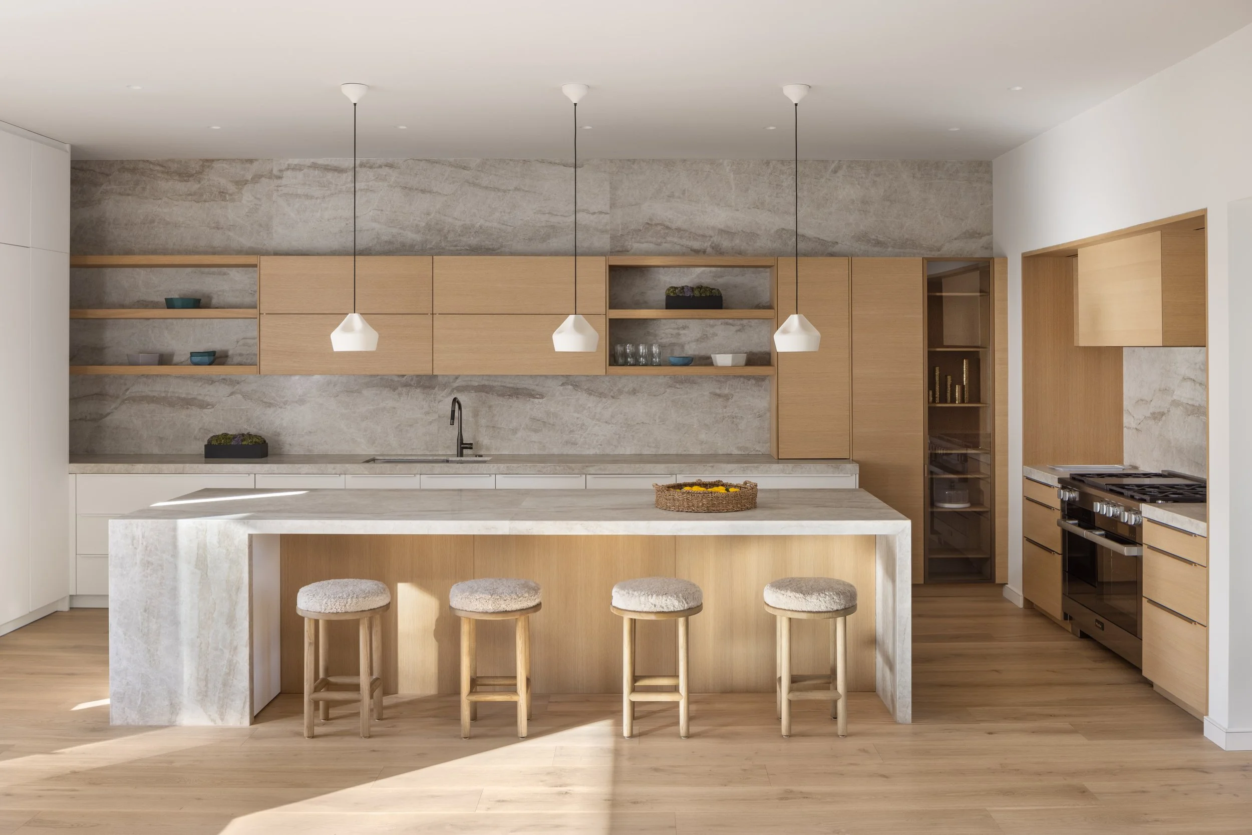 Modern kitchen with wooden cabinets, a marble island, open shelves with dishes, three pendant lights, and a gray stone backsplash.