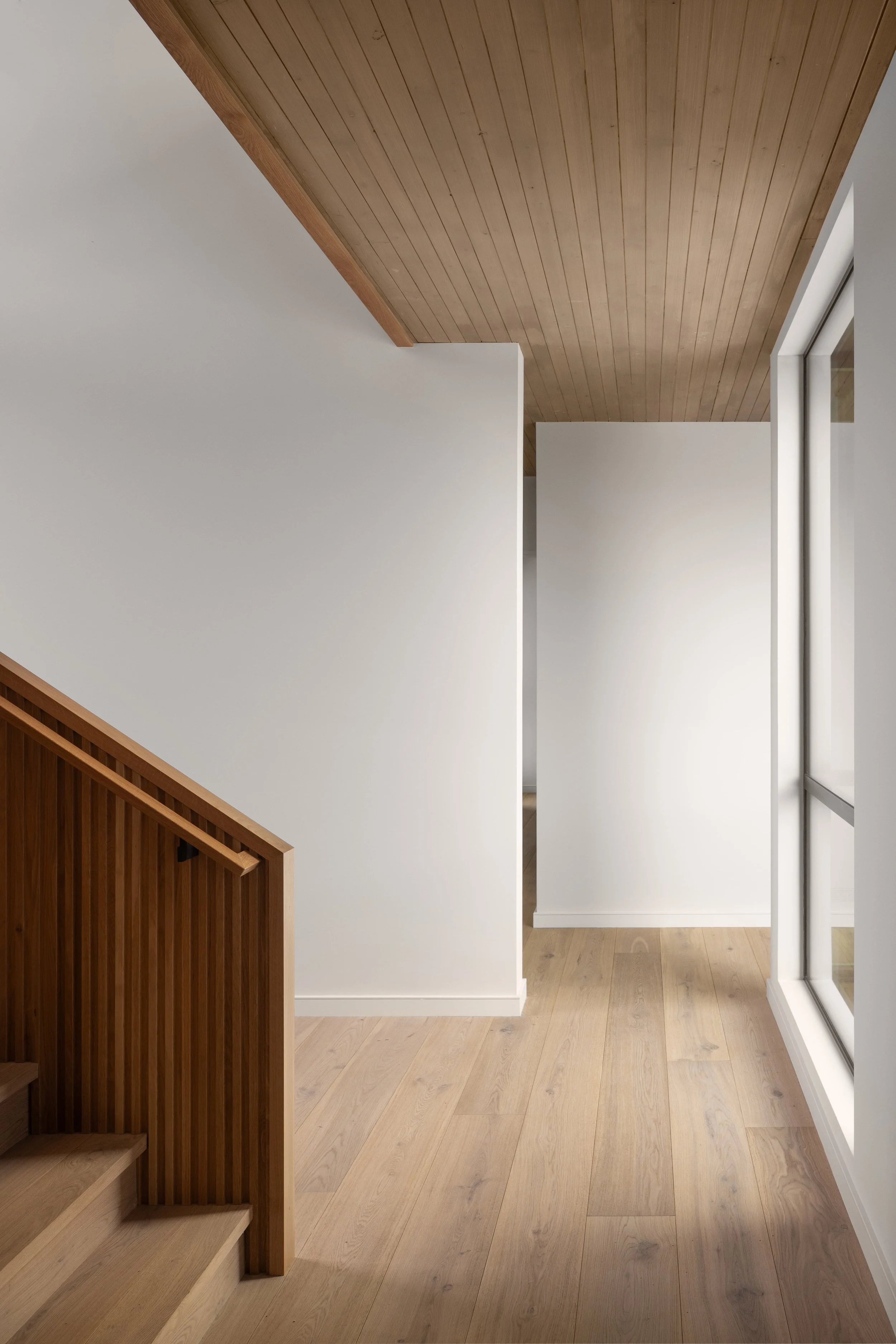Teton House in Jackson, Wyoming. Interior view of a modern staircase with a wooden handrail and a white wall, natural light, and large windows in a contemporary home.