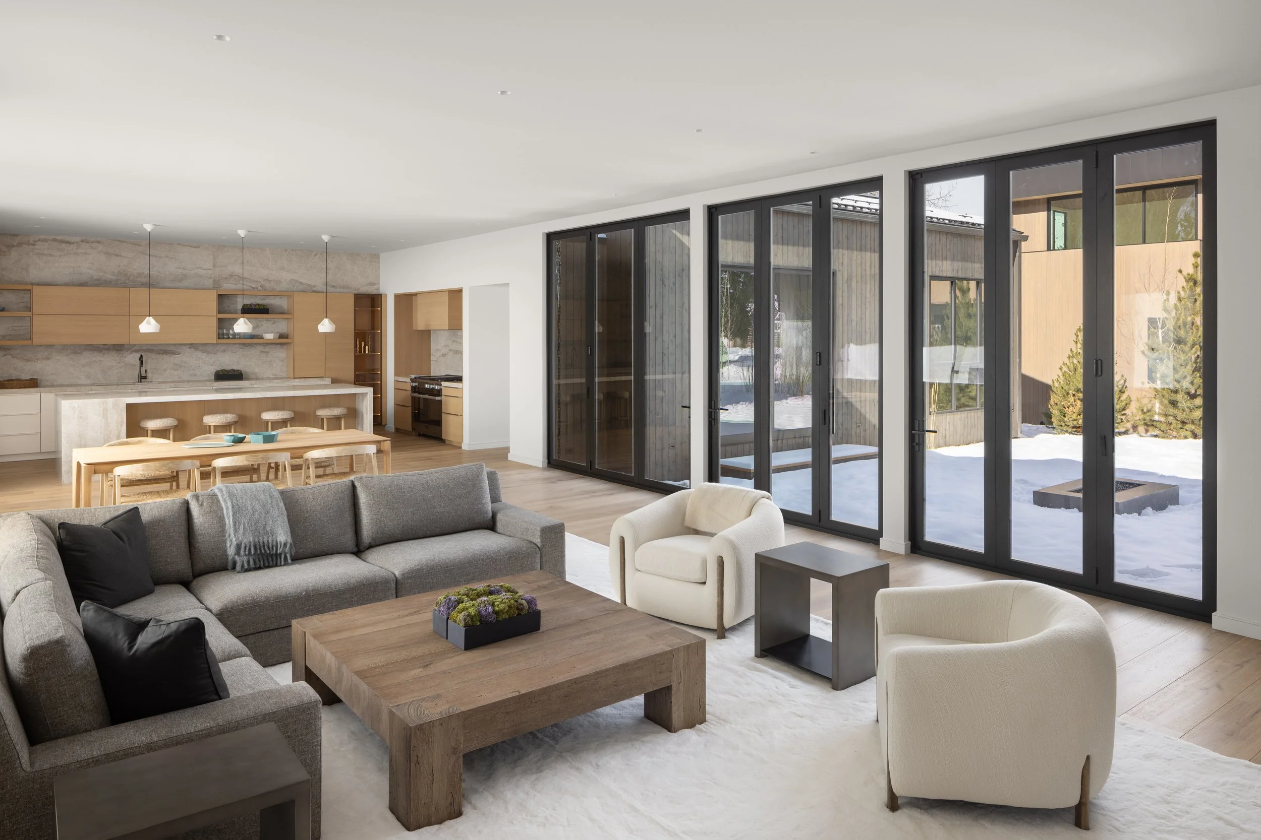 Teton House in Jackson, Wyoming. Modern living room with large floor-to-ceiling windows showing snow outside, grey sectional sofa, white armchair, wooden coffee table, minimalist decor.