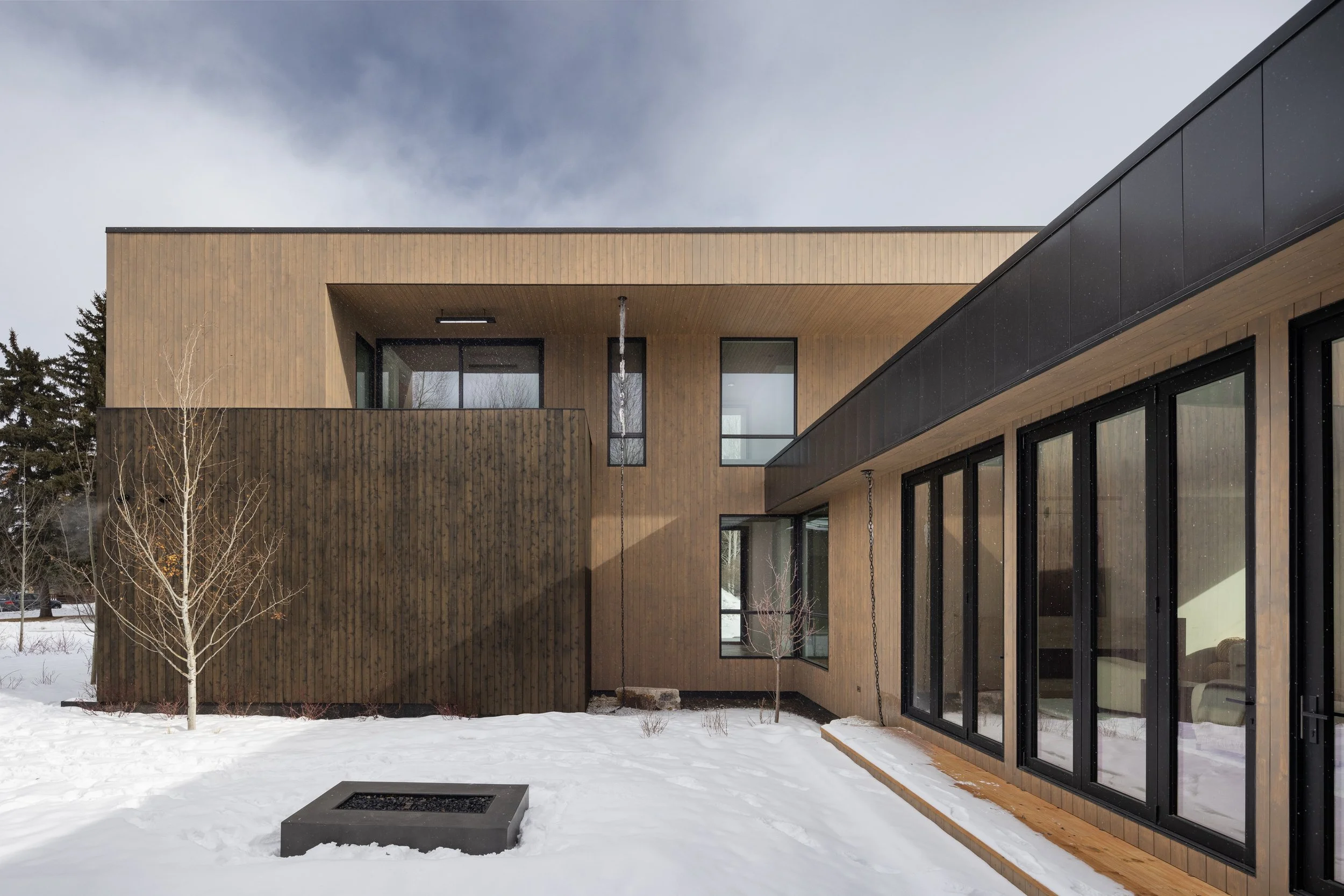 Teton House in Jackson, Wyoming. Modern wooden house with large glass windows, situated in a snowy landscape with leafless trees and a fire pit in the foreground.