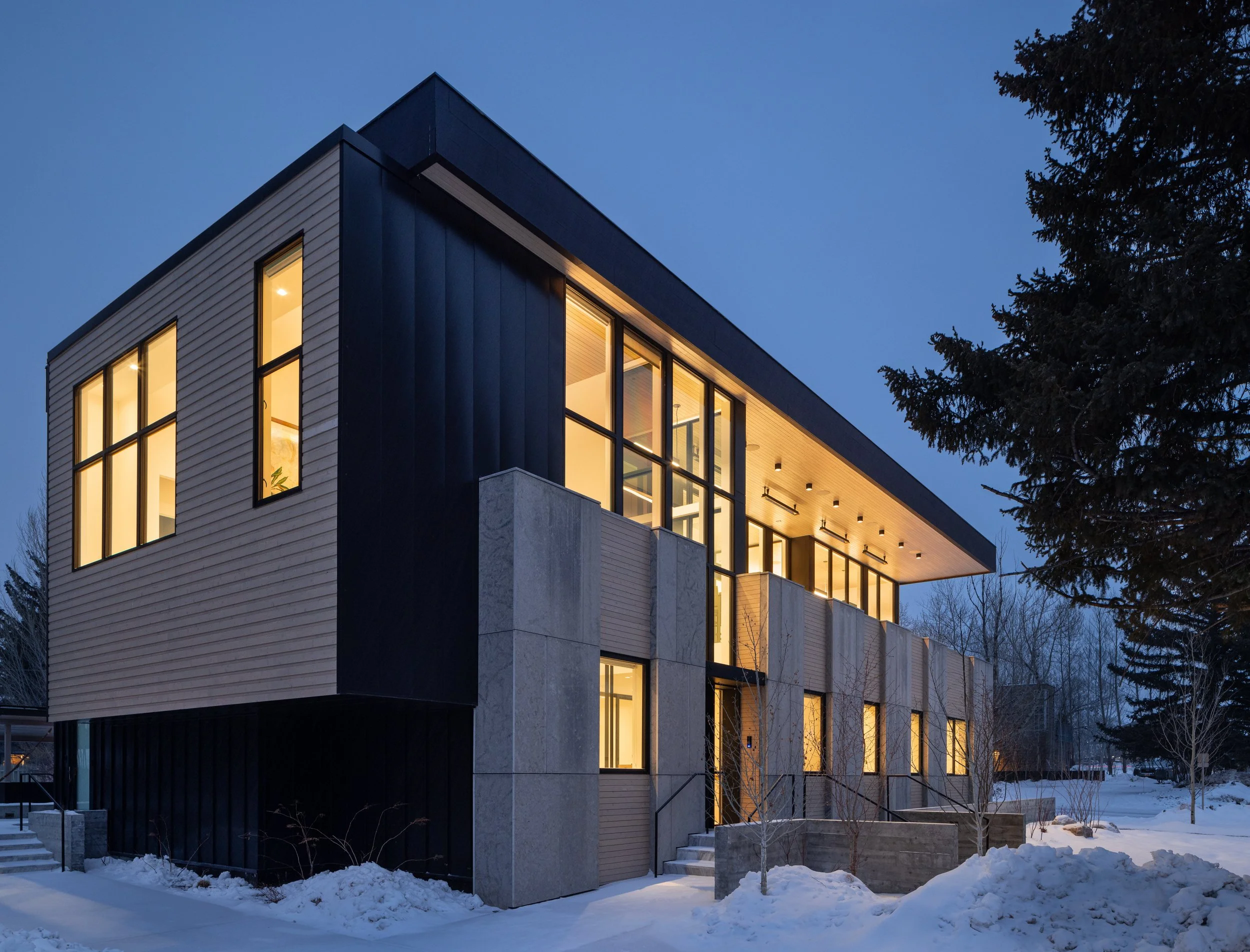 Modern multi-story house with large windows illuminated from inside, set in a snowy landscape with bare trees and a large evergreen tree nearby in the evening.