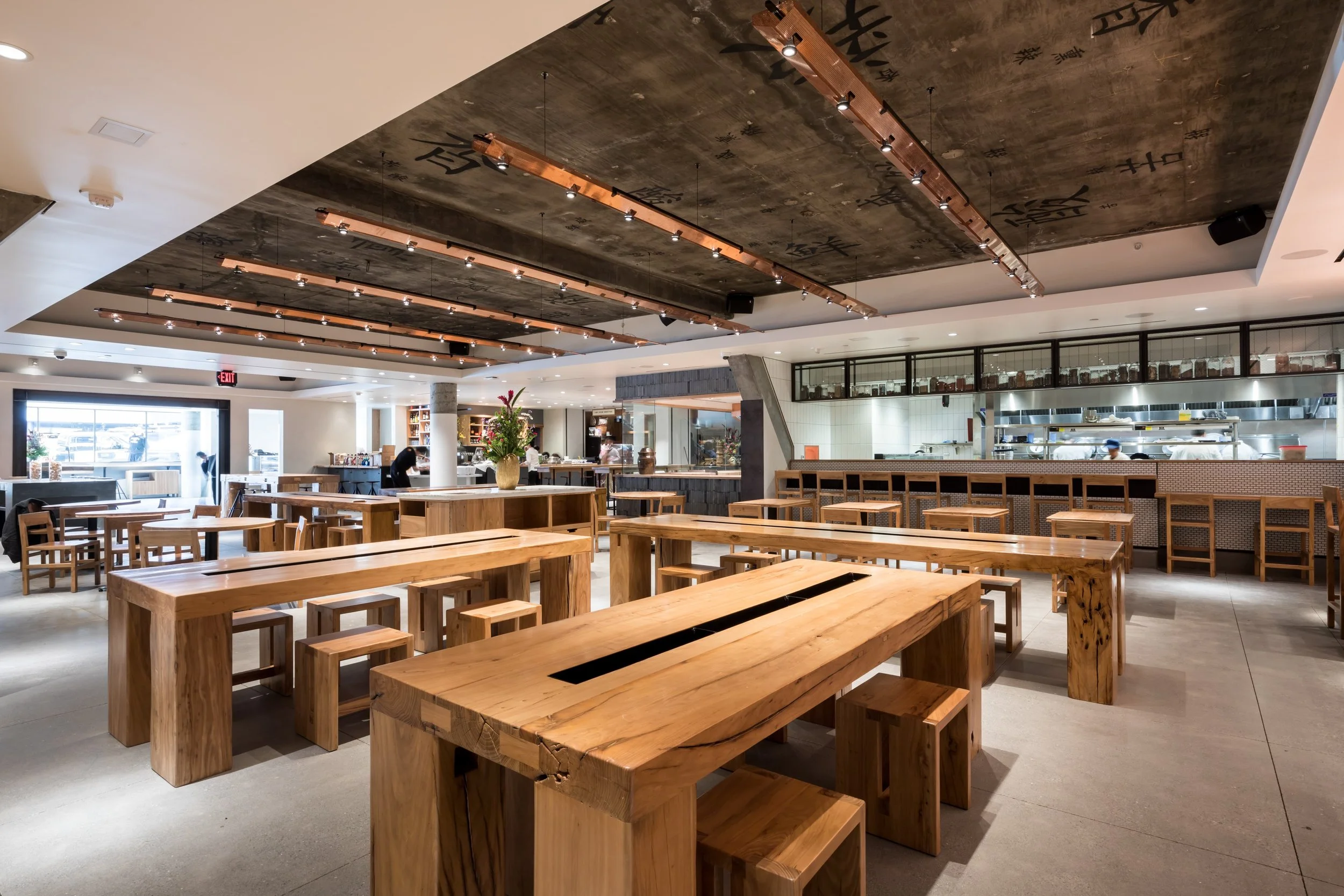 A spacious restaurant interior with wooden tables and chairs, a decorated counter, and an open kitchen area visible in the background. The ceiling features hanging wooden lights with Chinese characters painted on it.