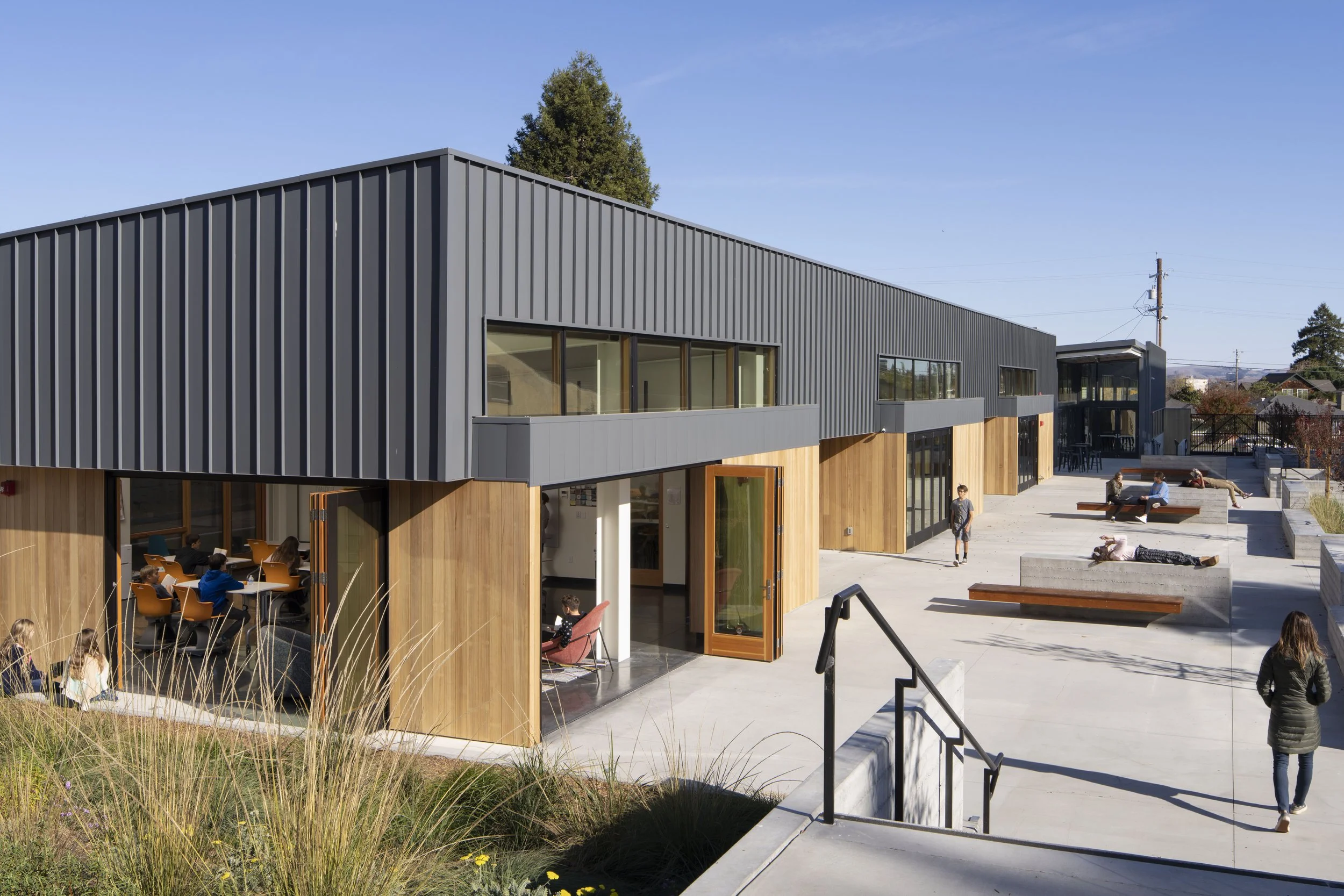 Modern school building with a gray metal exterior and large windows, surrounded by an outdoor courtyard where students are sitting and walking on a sunny day.