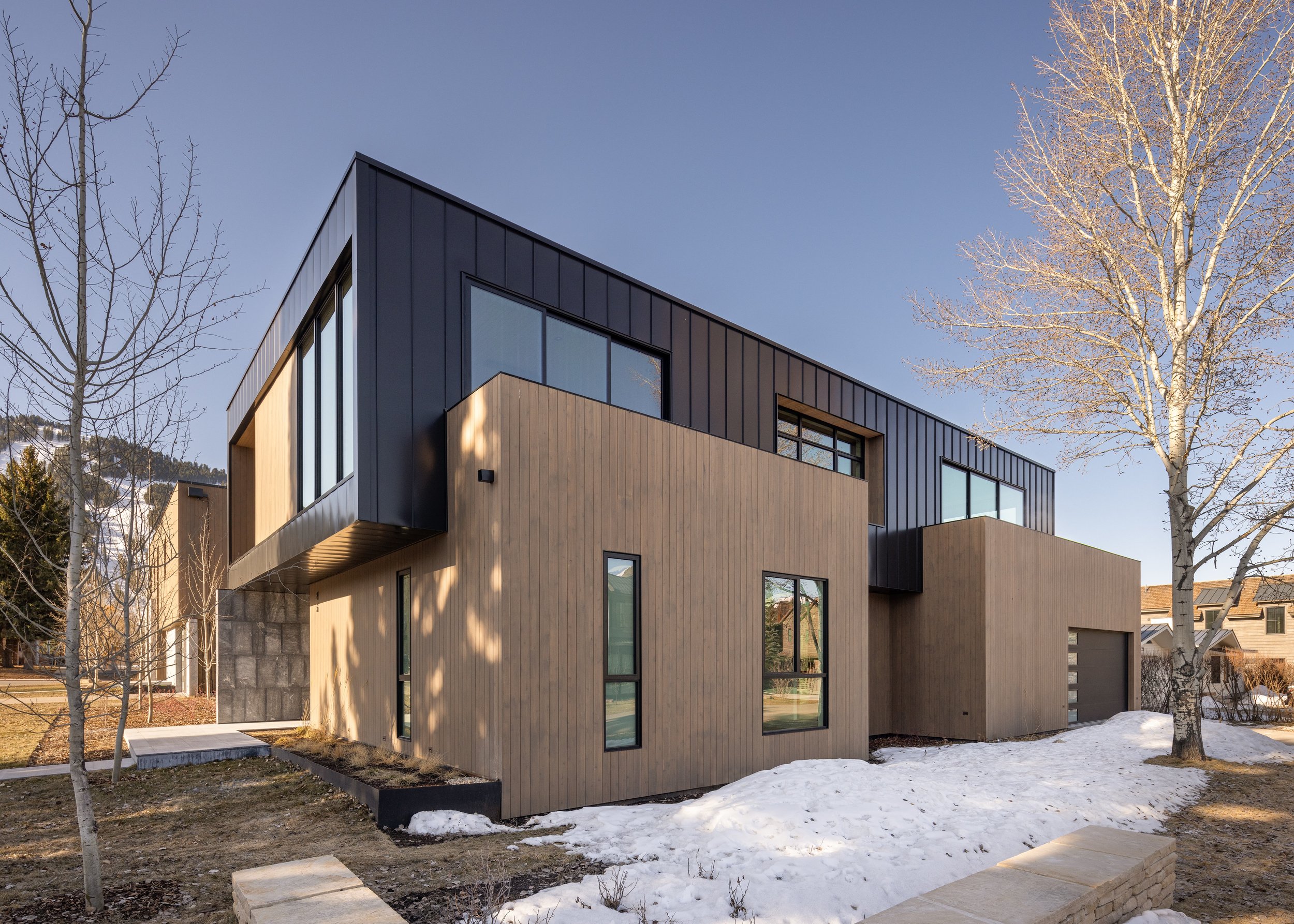 Split Timber House in Jackson, Wyoming. Modern multi-story residential building with black and wooden exterior, large windows, surrounded by leafless trees, patches of snow on the ground, under a clear blue sky.