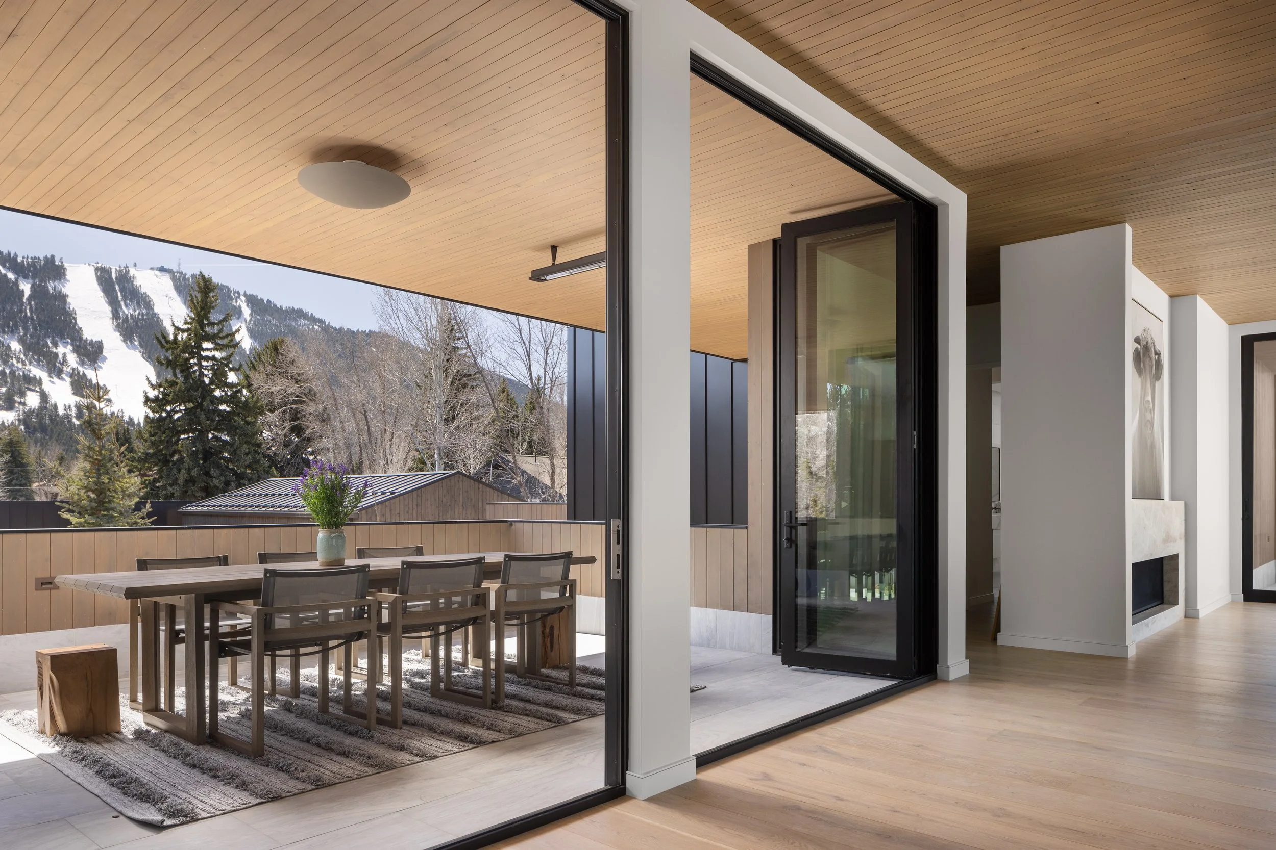 Split Timber House in Jackson, Wyoming. Modern indoor-outdoor living space with a wooden ceiling, glass sliding doors, an outdoor dining area with a wooden table and chairs, with snowy mountain scenery in the background.
