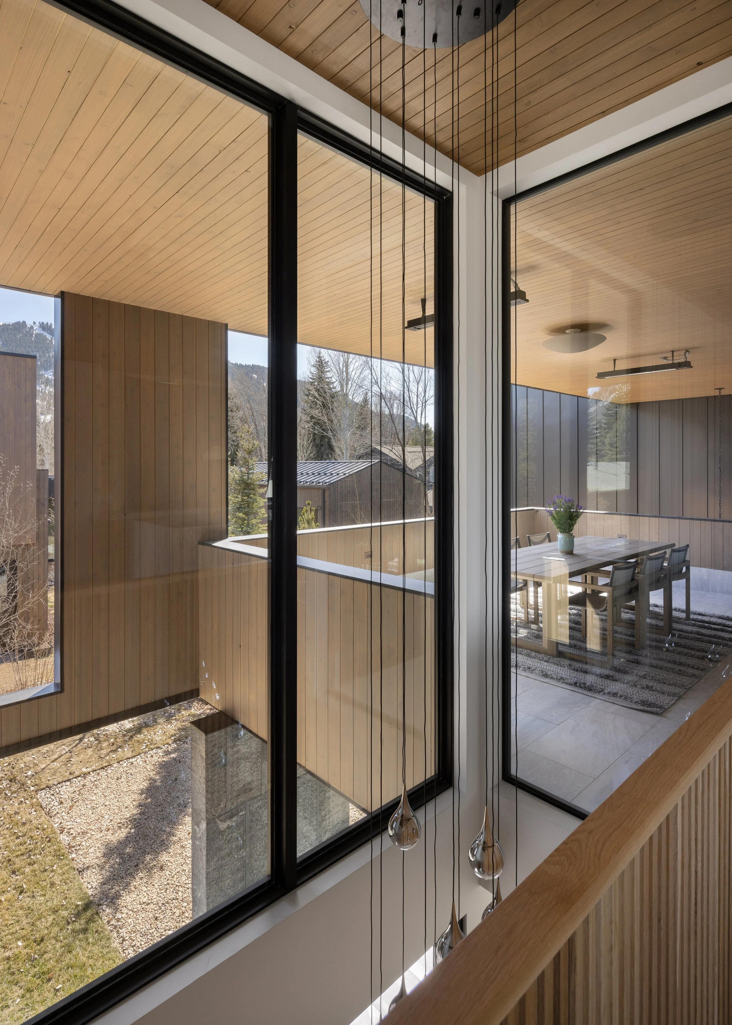 Split Timber House in Jackson, Wyoming. Interior view of a modern house with large glass windows, wooden ceiling, and a dining area with a vase of flowers on a table.