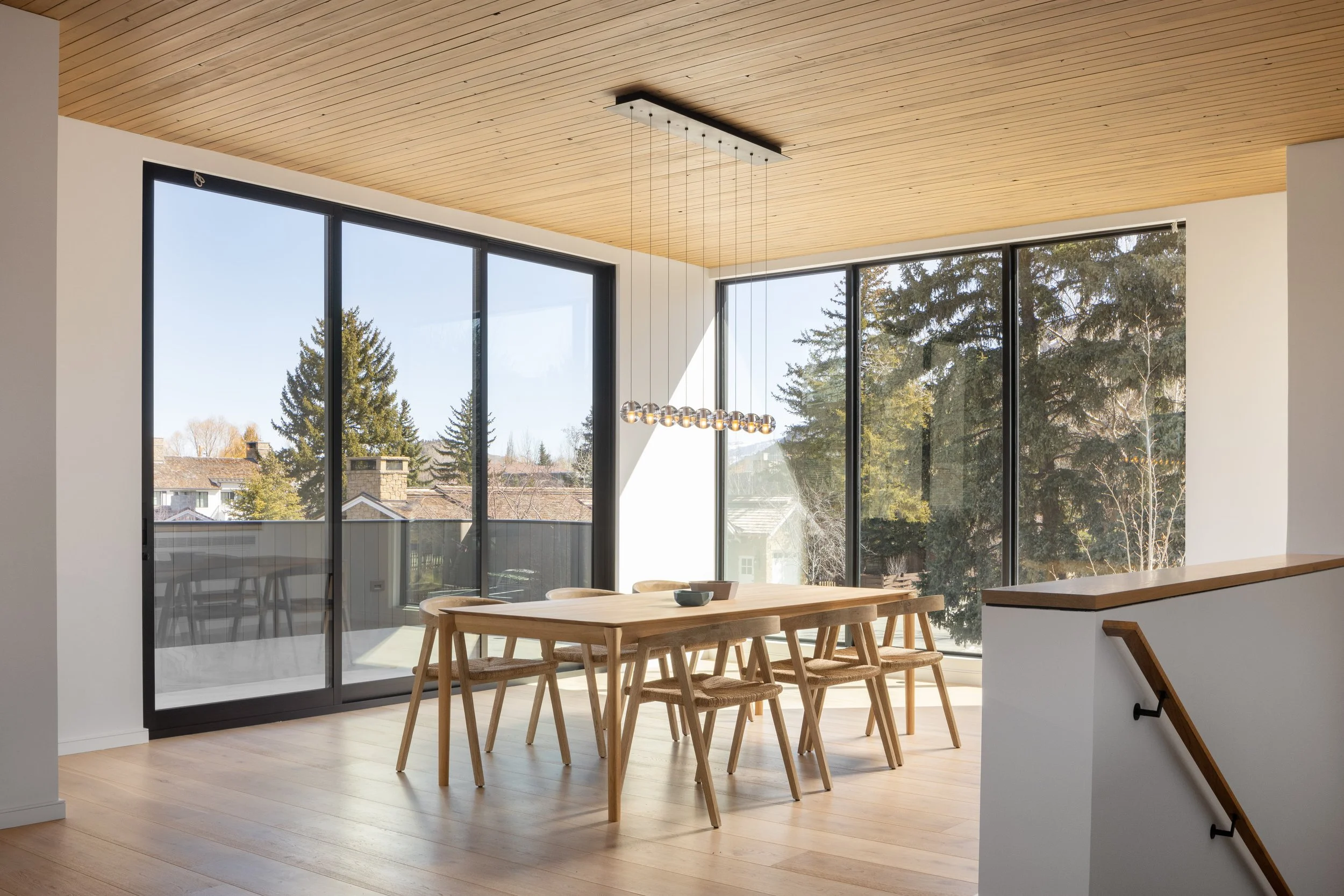 Split Timber House in Jackson, Wyoming. Bright dining area with large floor-to-ceiling windows, wooden ceiling, minimalist wooden dining table, and chairs, overlooking trees and a neighborhood.