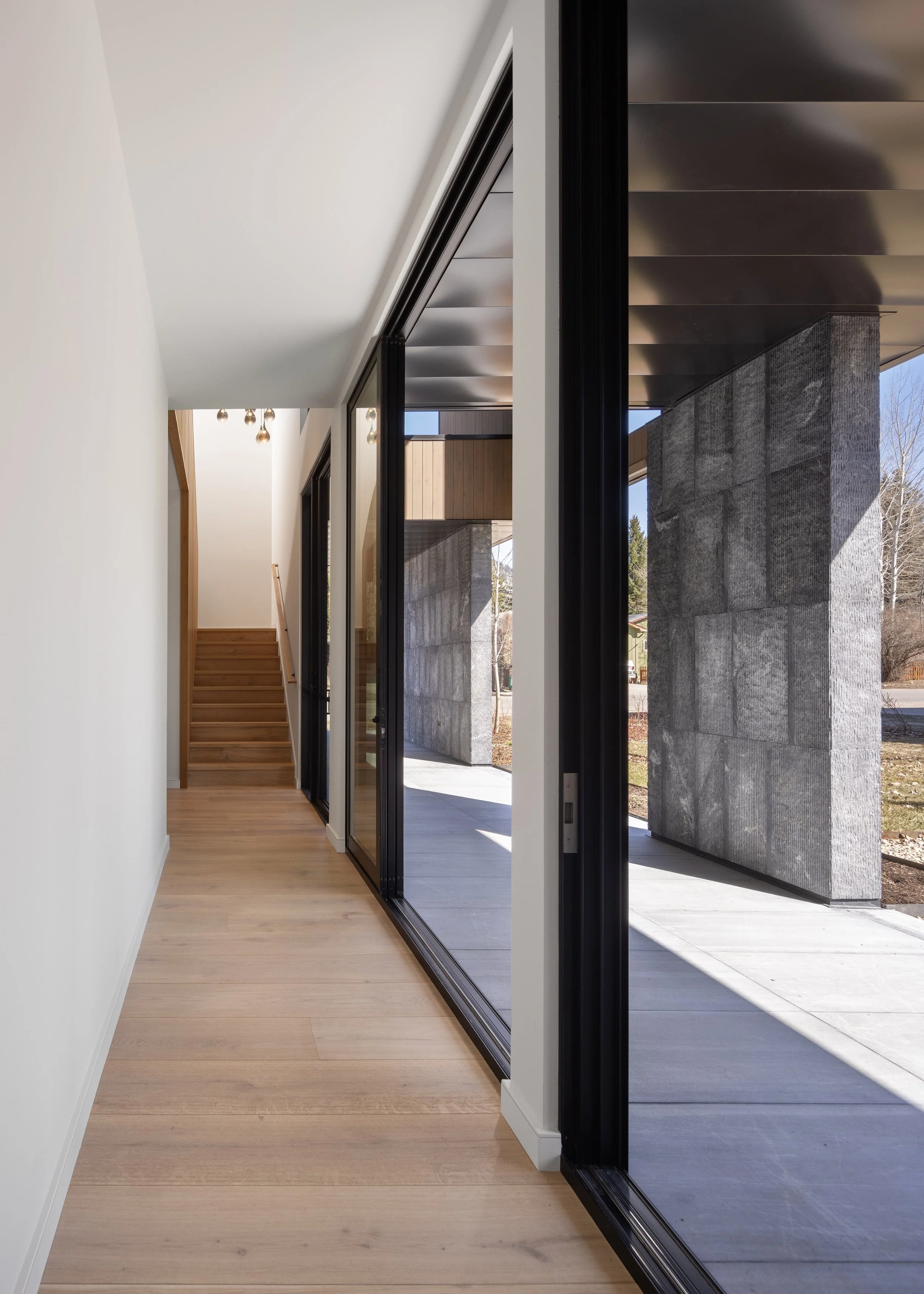 Split Timber House in Jackson, Wyoming. Modern interior hallway with wooden flooring, white walls, and large glass sliding doors leading to an outdoor area with stone pillars and a pergola.