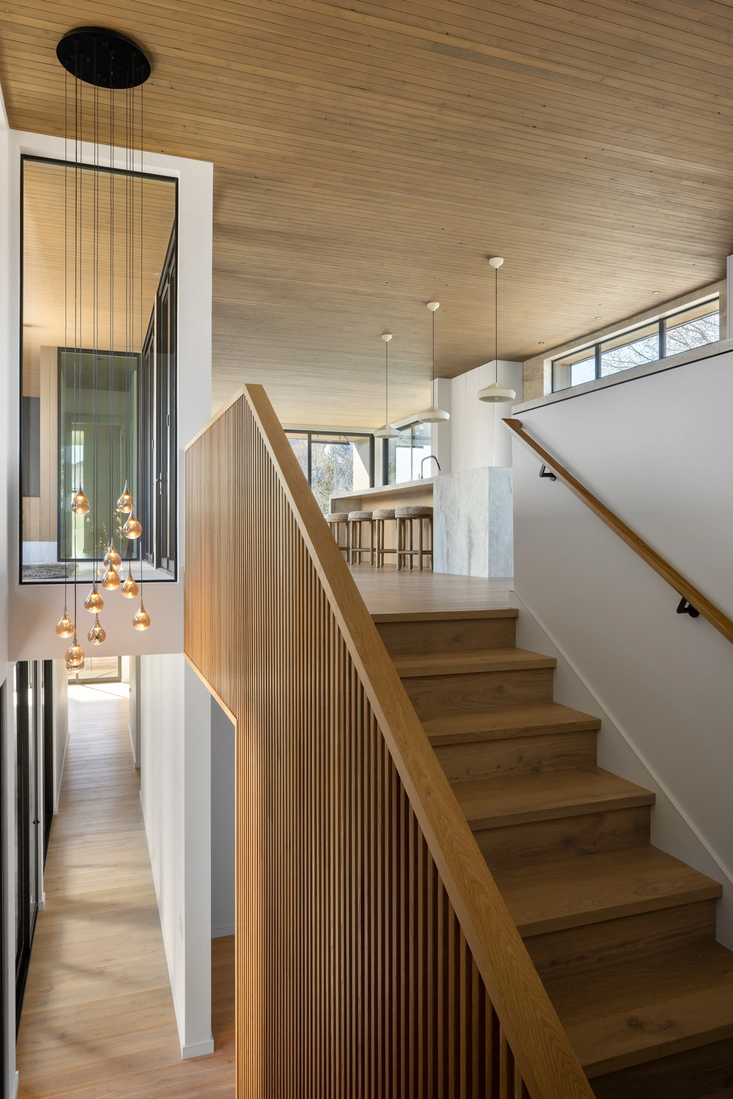 Split Timber House in Jackson, Wyoming. Interior of a modern home with wooden stairs, a wooden ceiling, and large windows, featuring minimalist pendant lights and a white kitchen island in the background.
