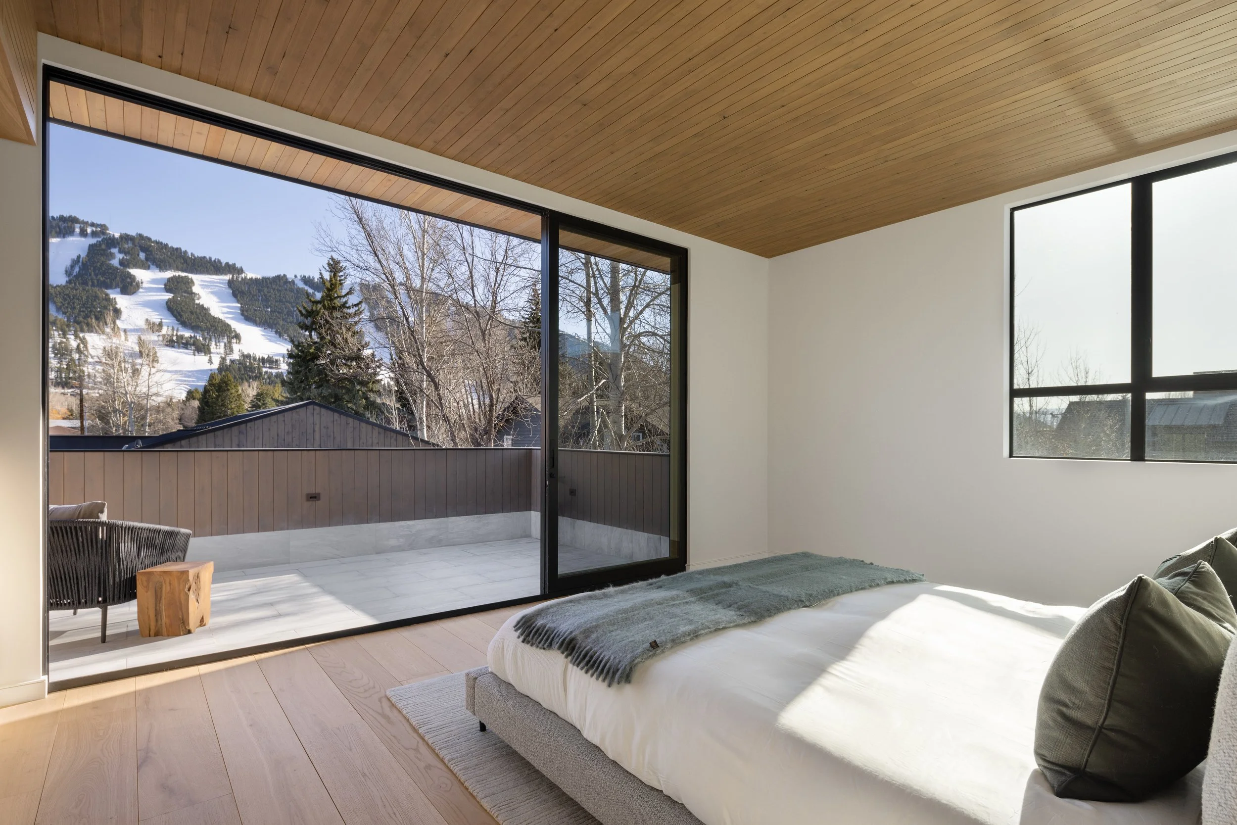 Split Timber House in Jackson, Wyoming. Modern bedroom with large glass doors opening to a mountain view, light wood ceiling, white walls, and a neutral color bed with gray pillows.