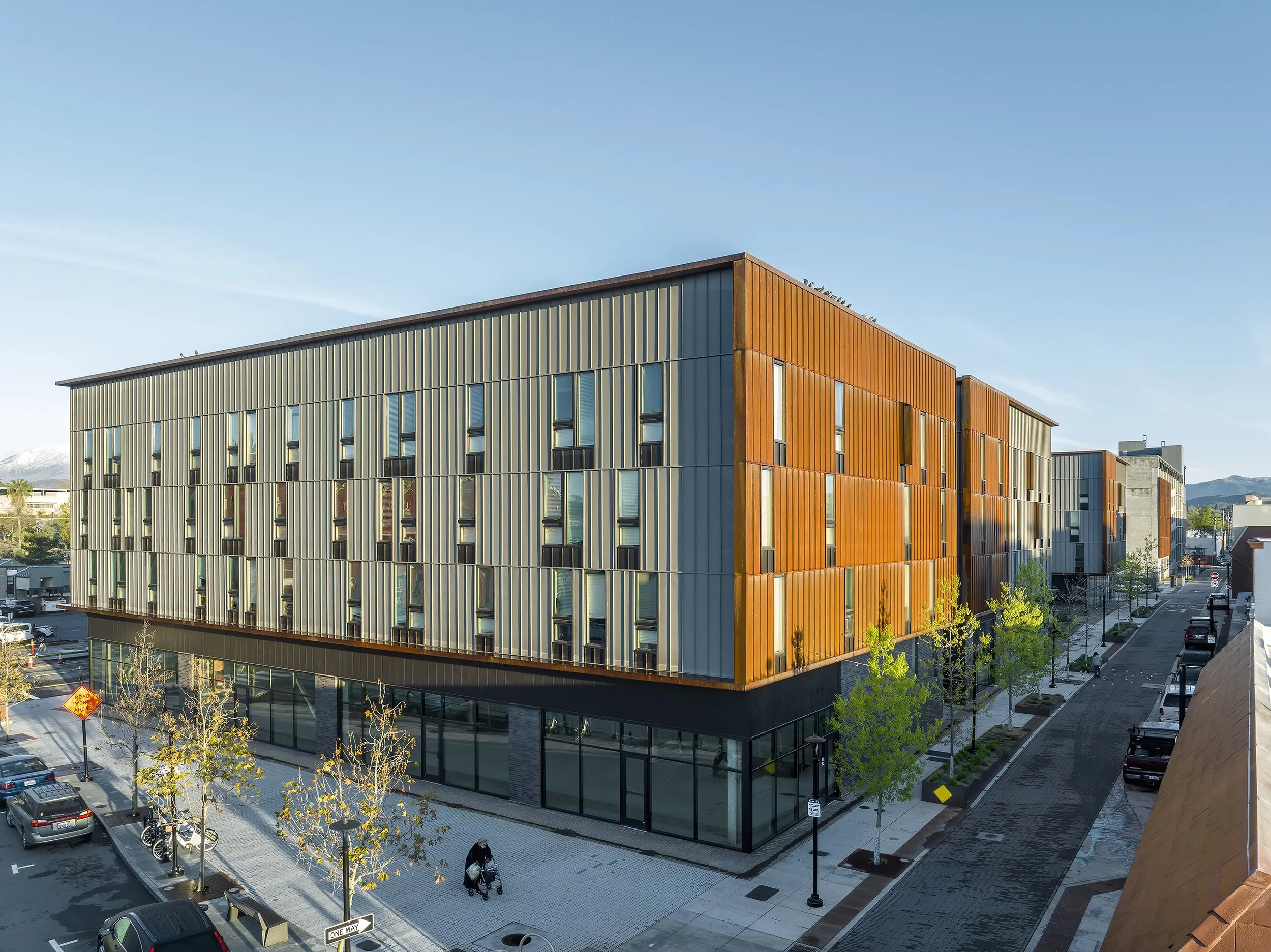 Modern multi-story building with metal siding in bronze and beige tones, large glass windows on the ground floor, and a clear blue sky above. Surrounding area includes parked cars, young trees, and a sidewalk with a pedestrian pushing a stroller.