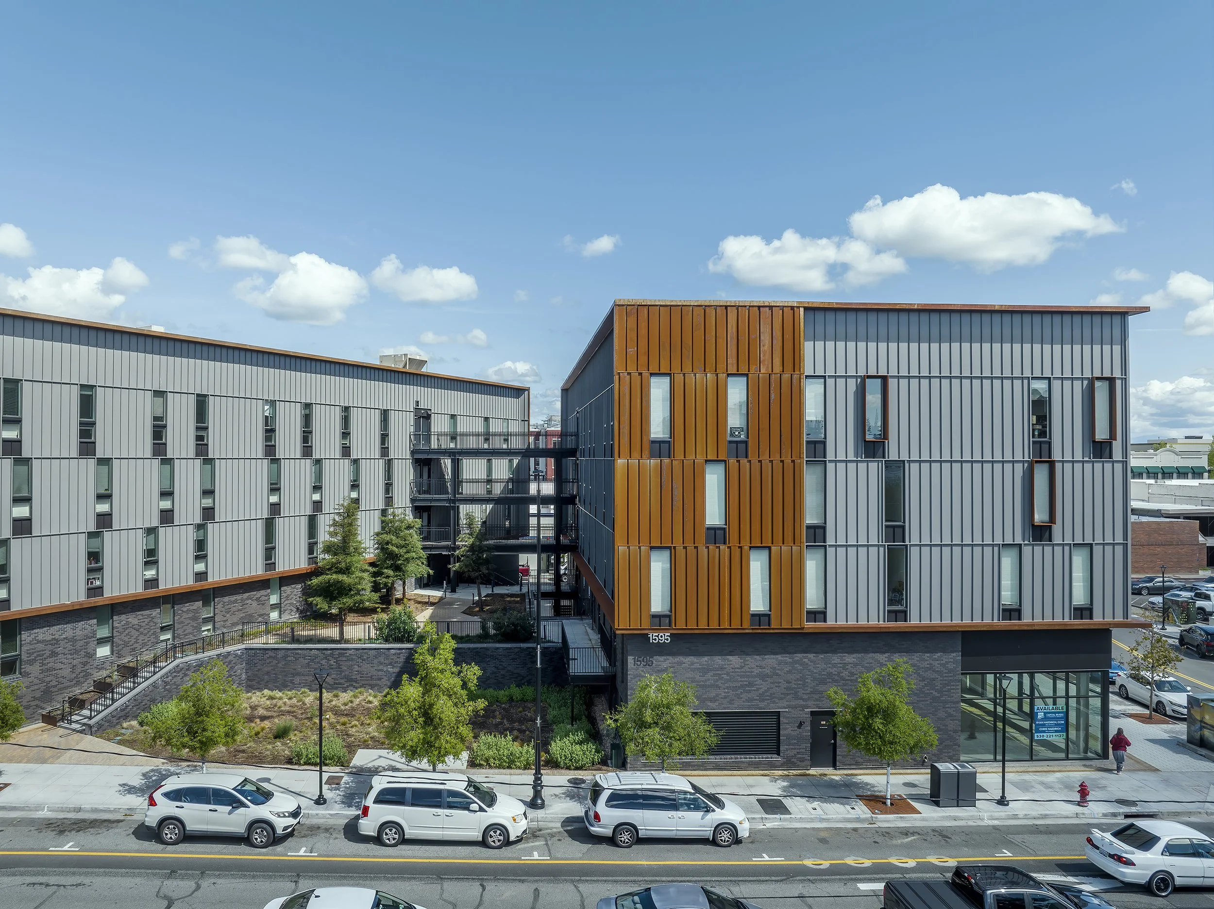Block 7 Development in Redding, California. California Place affordable housing. Modern multi-story residential and commercial buildings with gray and orange exterior, parking lot, and a sidewalk with pedestrians, under a blue sky with clouds.