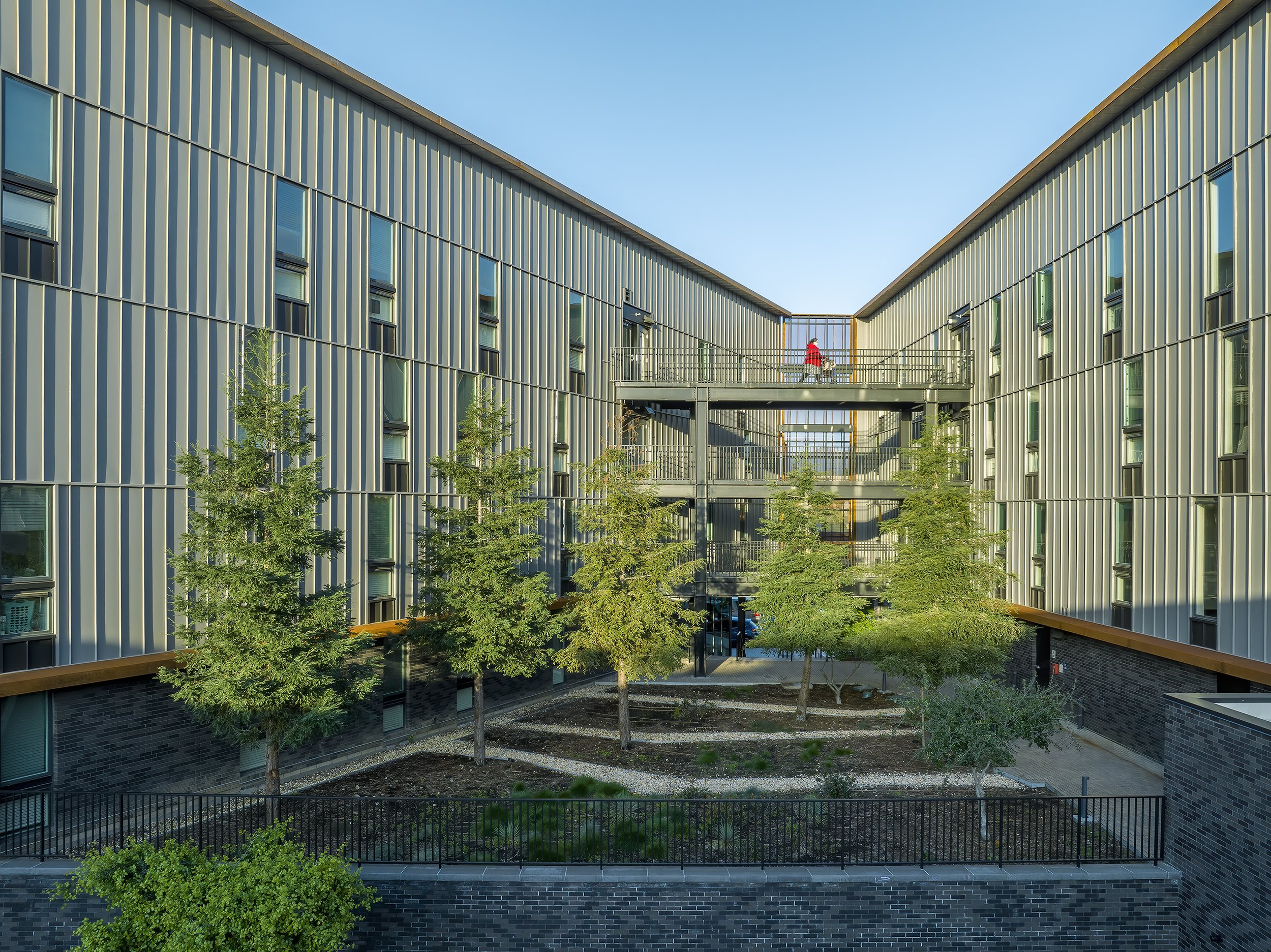 Block 7 Development in Redding, California. California Place affordable housing. Modern apartment building with vertical siding, surrounded by greenery, with an open courtyard and walkway connecting the buildings. The sky is clear and blue.