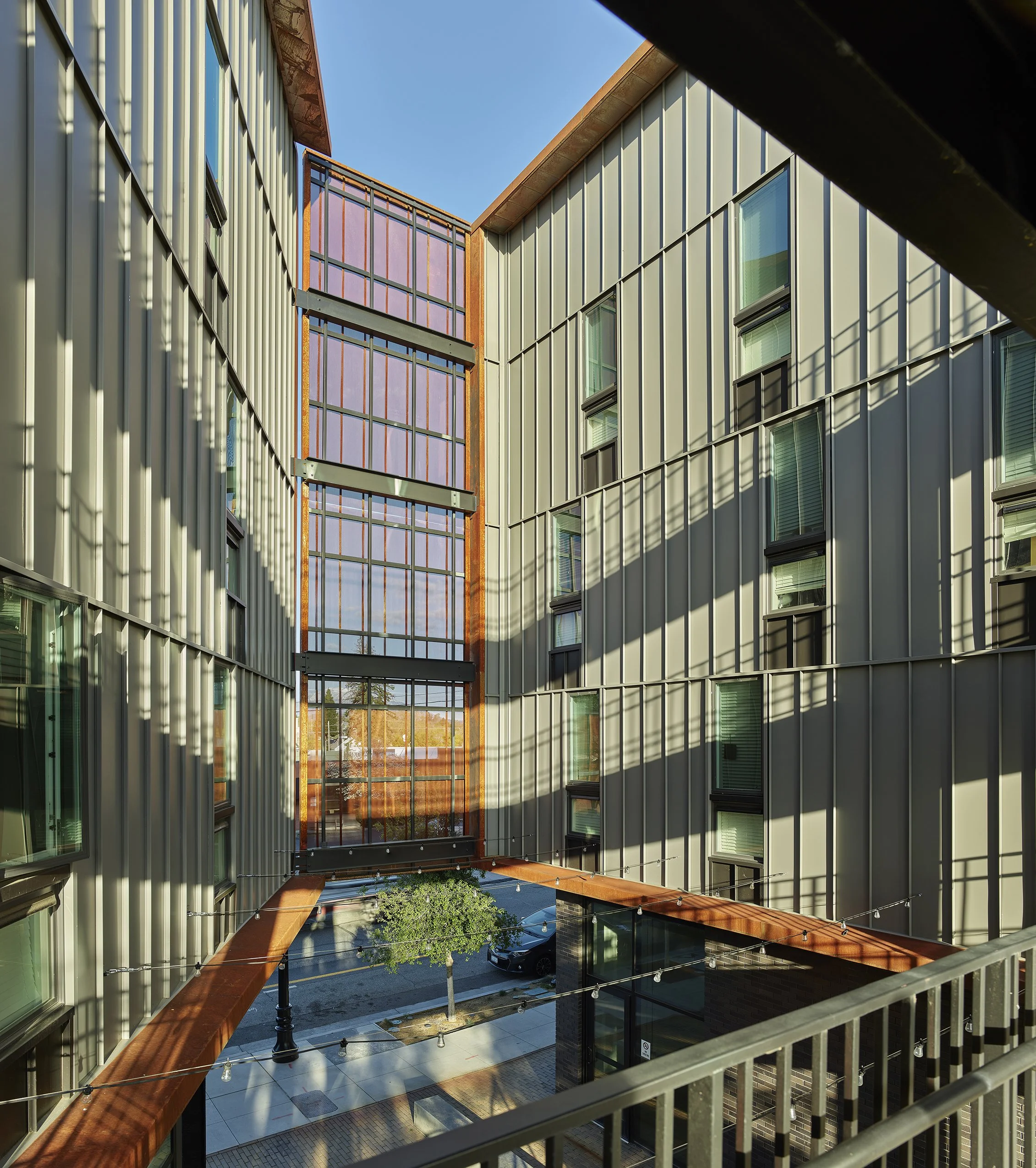 Block 7 Development in Redding, California. California Place affordable housing. View of a modern building courtyard with metal siding, large windows, string lights, trees, and a parking lot with a car, under a clear blue sky.