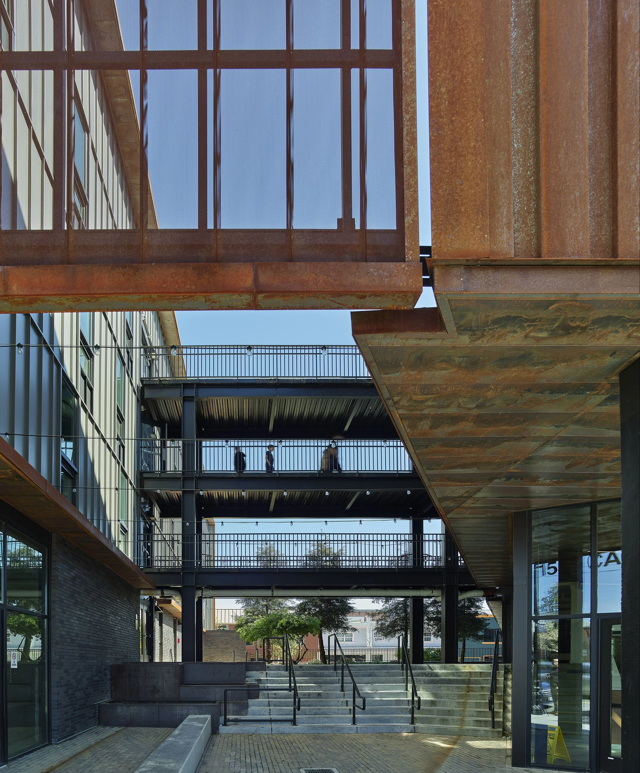 Block 7 Development in Redding, California. California Place affordable housing. An outdoor view of a modern building with multiple levels and stairs, featuring glass windows, metal railings, and a brick wall, with trees.