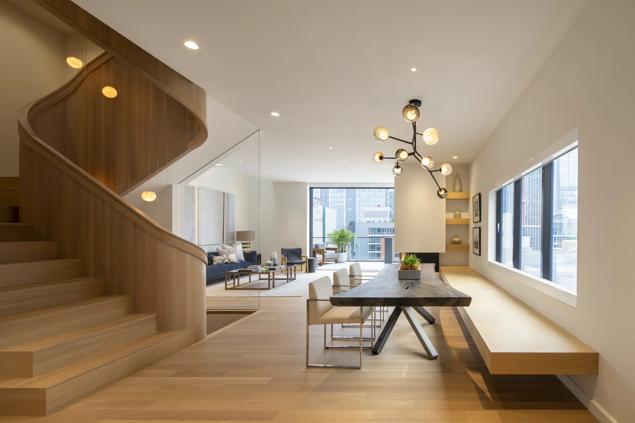 Modern open-concept living and dining area with wooden flooring and a staircase on the left, featuring a large window and contemporary lighting fixtures.