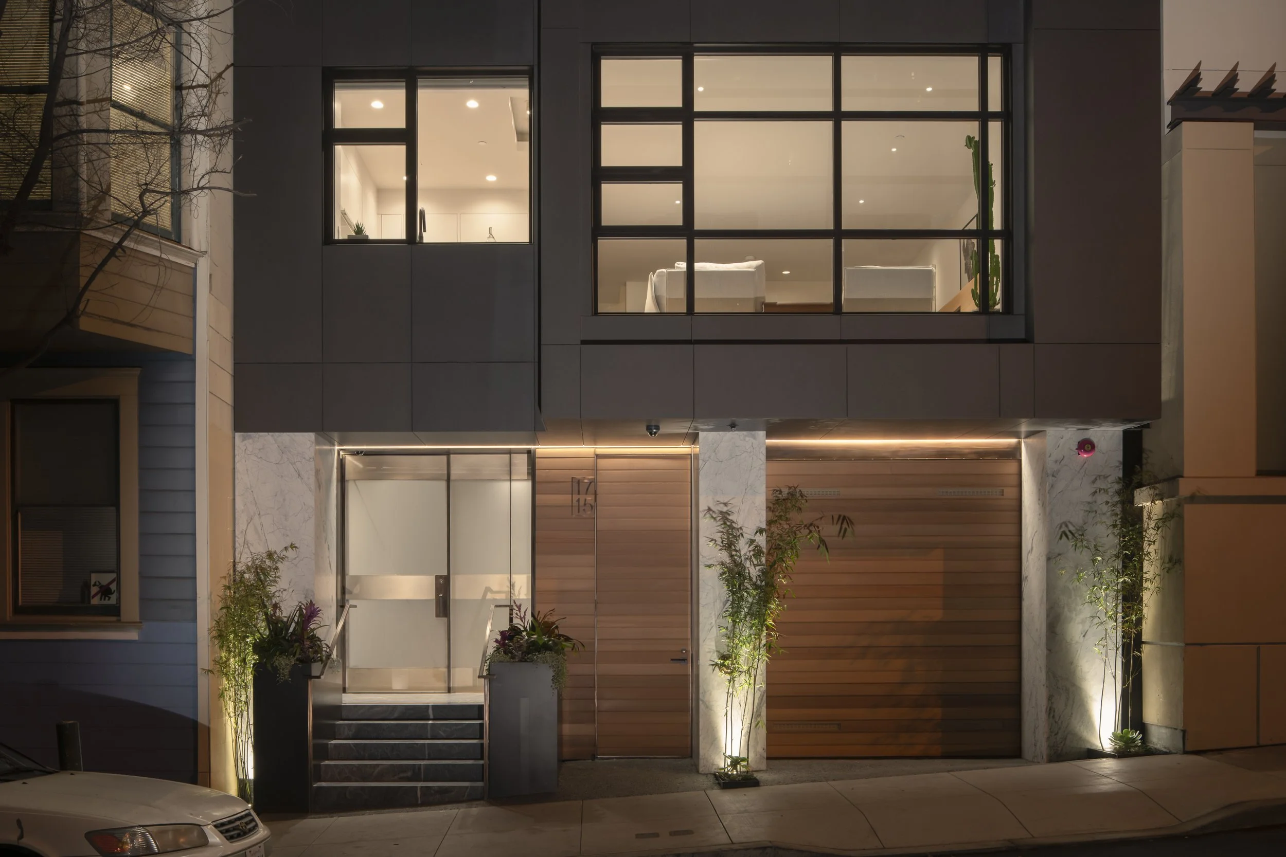 Six-Story-City House in San Francisco, California. Modern multi-story house with large glass windows, a garage with wooden door, and landscaped front yard with plants, illuminated at night.