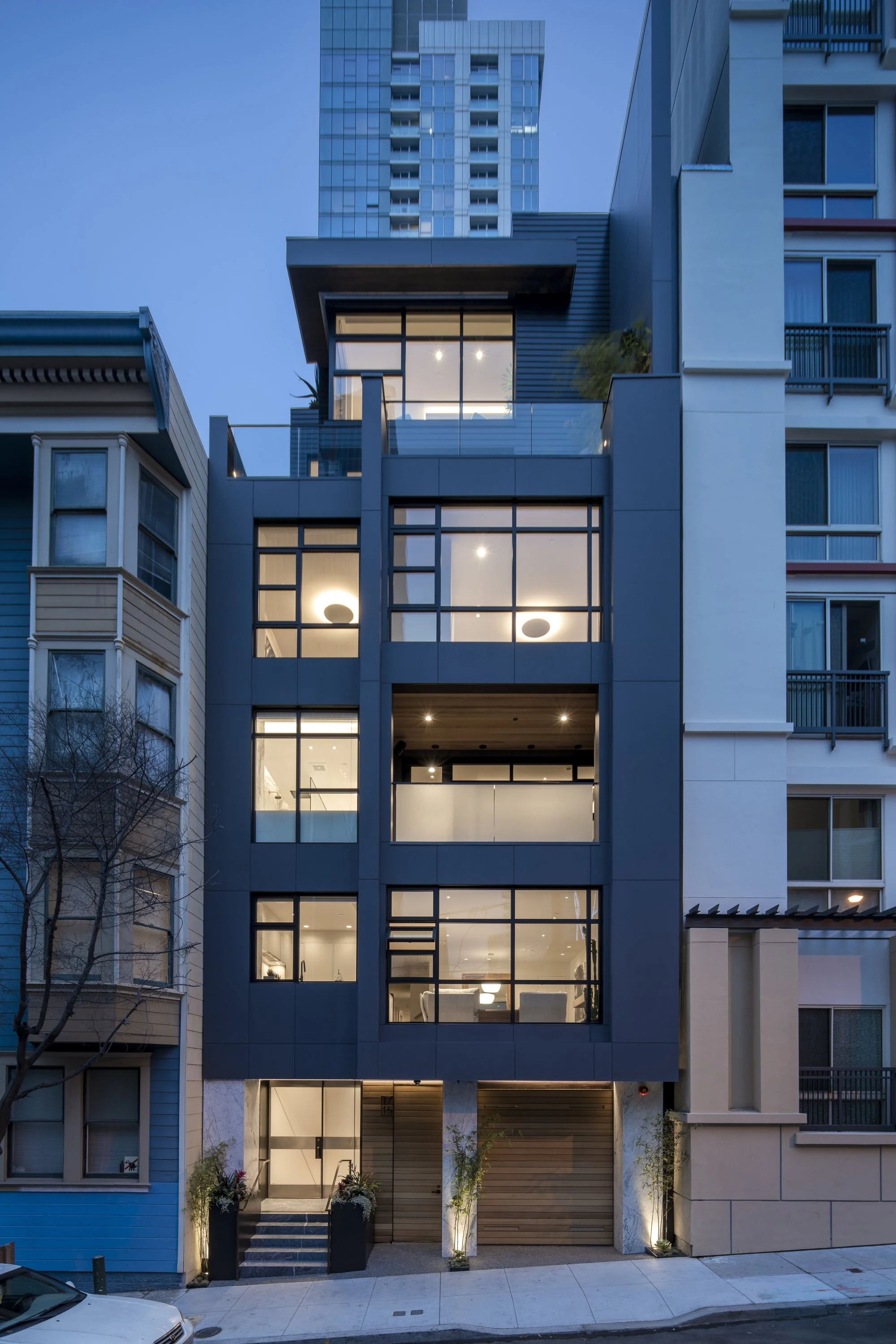 Six-Story-City House in San Francisco, California. Modern urban building with large glass windows, with some interior lighting visible, and decorative plants at the entrance. Adjacent older buildings with traditional design on either side.