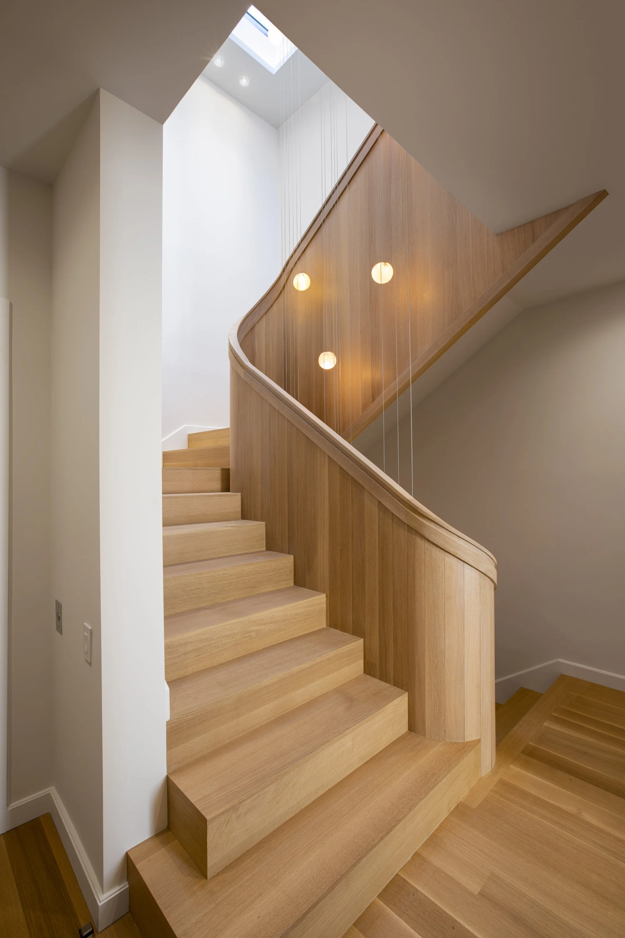 Six-Story-City House in San Francisco, California. Wooden staircase with a curved handrail and modern hanging lights, seen from below near a skylight.