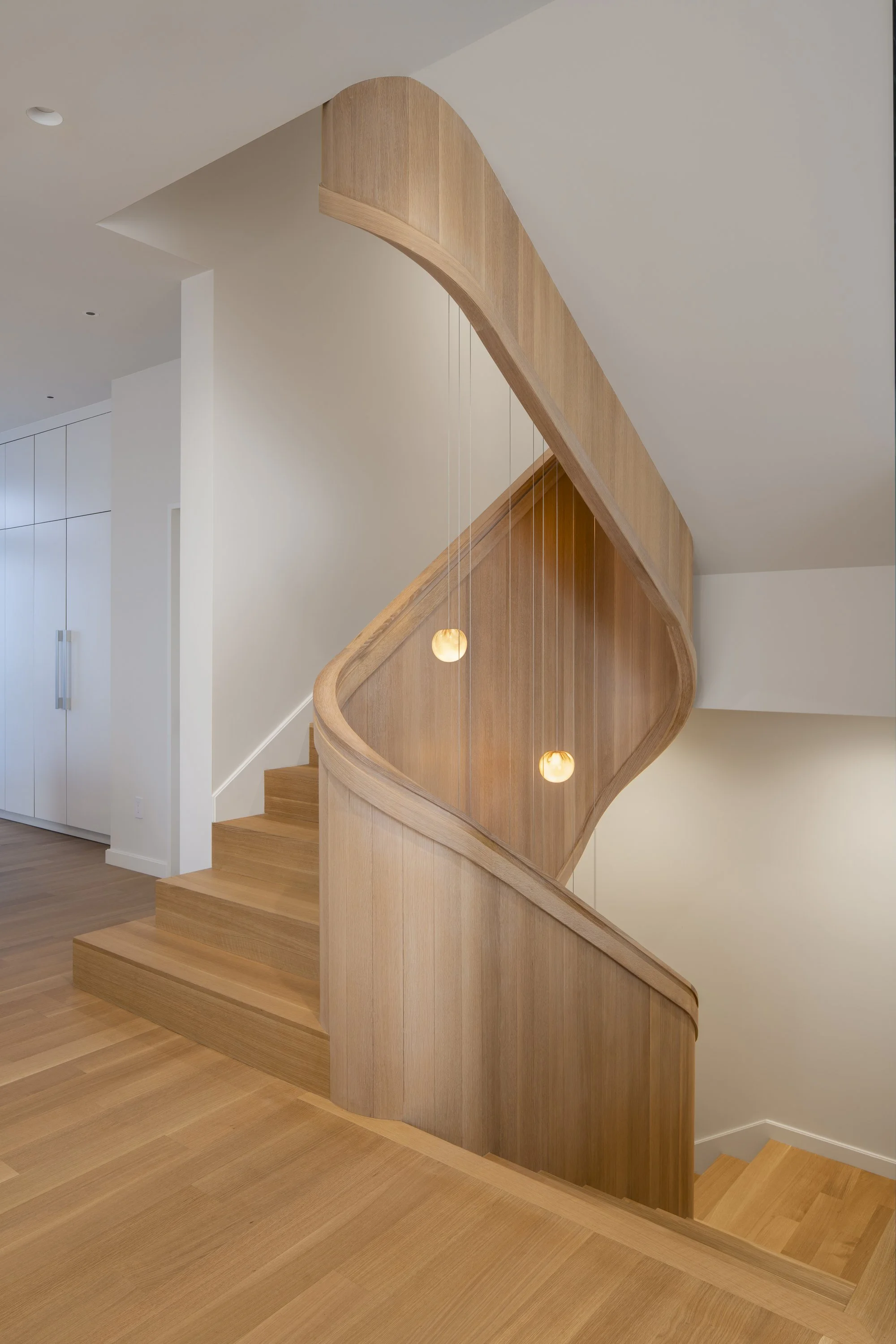 Six-Story-City House in San Francisco, California. Interior staircase with a curved wooden banister and steps, lit by hanging round lights, adjacent to white walls and cabinetry.