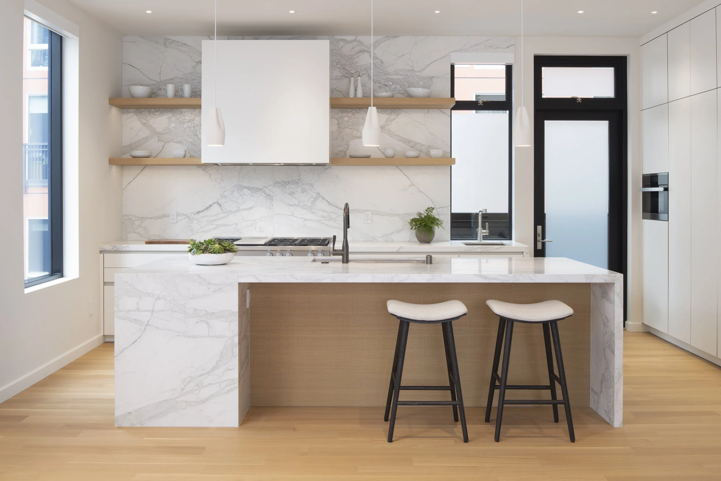Six-Story-City House in San Francisco, California. Modern kitchen with white marble island, two black bar stools, open shelving with white dishes, and black-framed window and door
