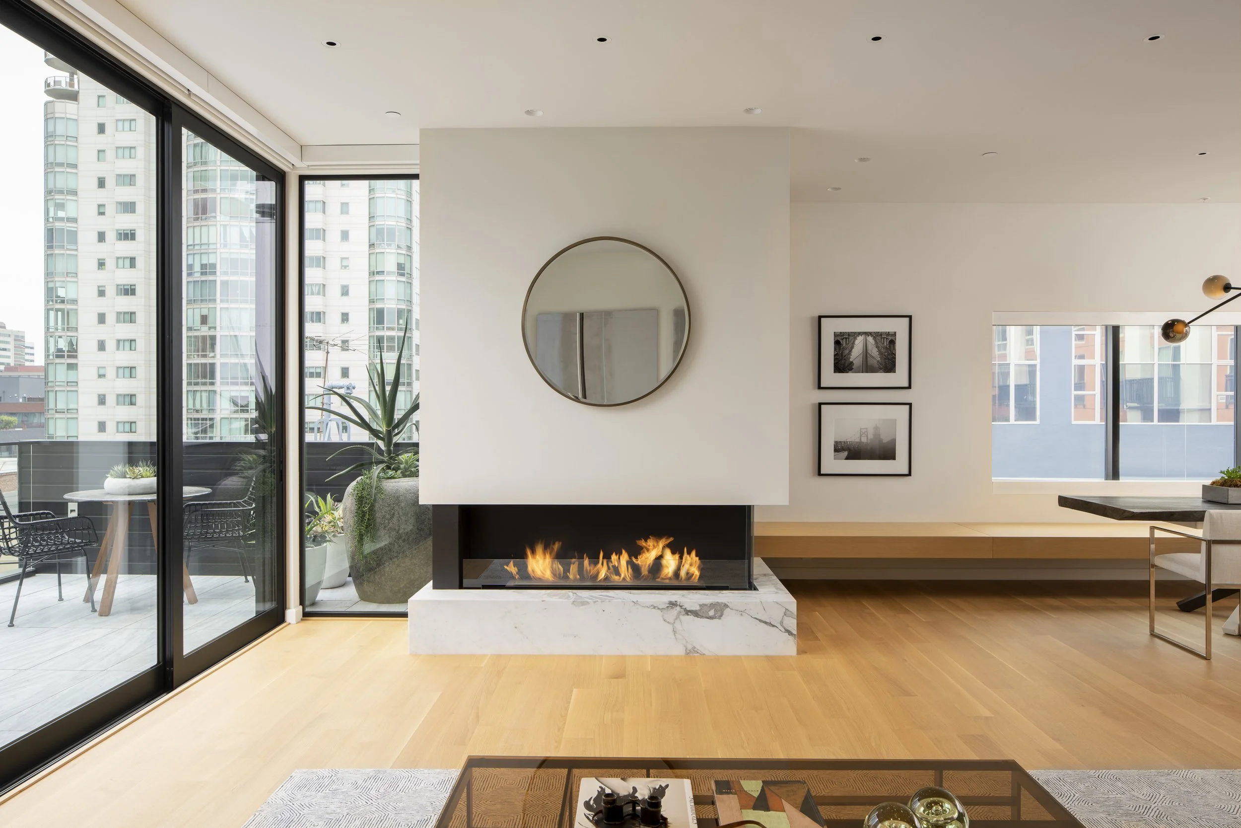 Six-Story-City House in San Francisco, California. Interior of a modern living room with a fireplace, large windows showing city buildings, wall art, and a balcony with outdoor seating.