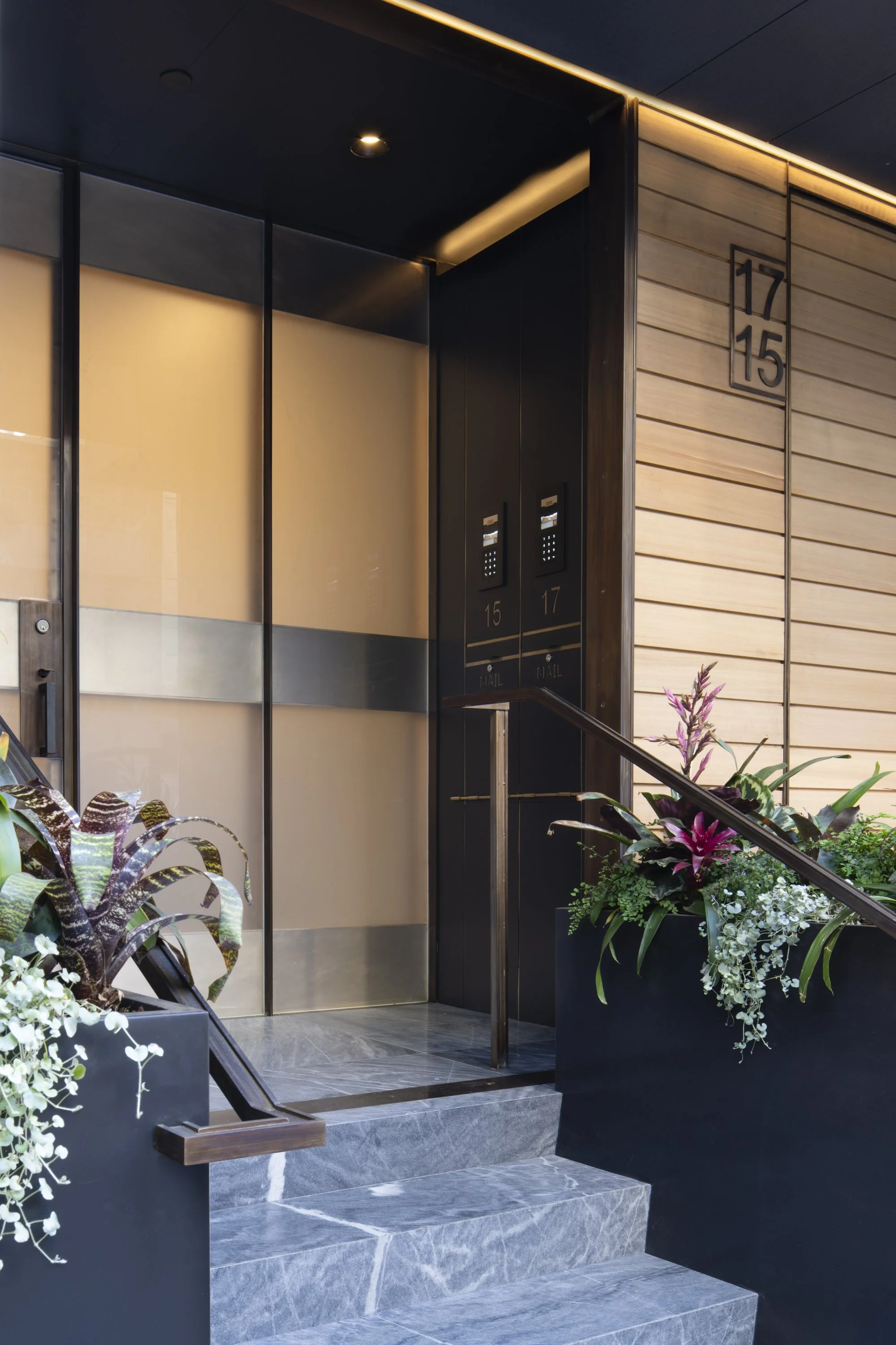 Six-Story-City House in San Francisco, California. Modern building entrance with elevator doors, house numbers 17 and 15 on a wood-paneled wall, and flower arrangements near the stairs.