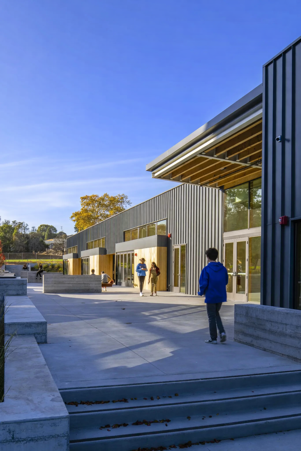 Spring Hill Montessori - Learning by Design in Petaluma, California. Students walking outside a modern school building with metal siding, large windows, and a concrete plaza, under a clear blue sky with some trees in the background.