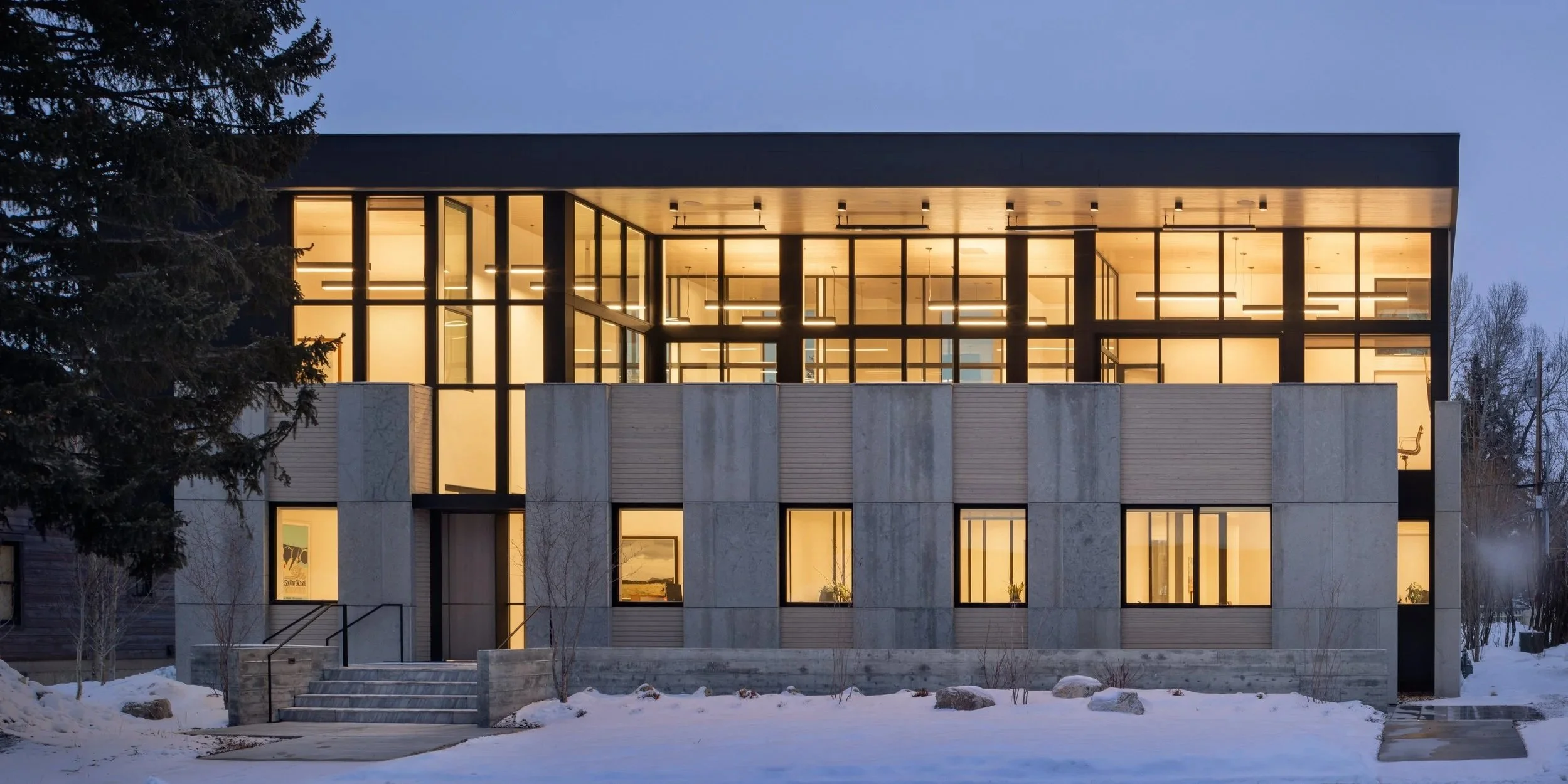 Office at Snow King in Jackson, Wyoming. Modern multi-story building with large glass windows, illuminated from inside, surrounded by snow, with a tree on the left side.