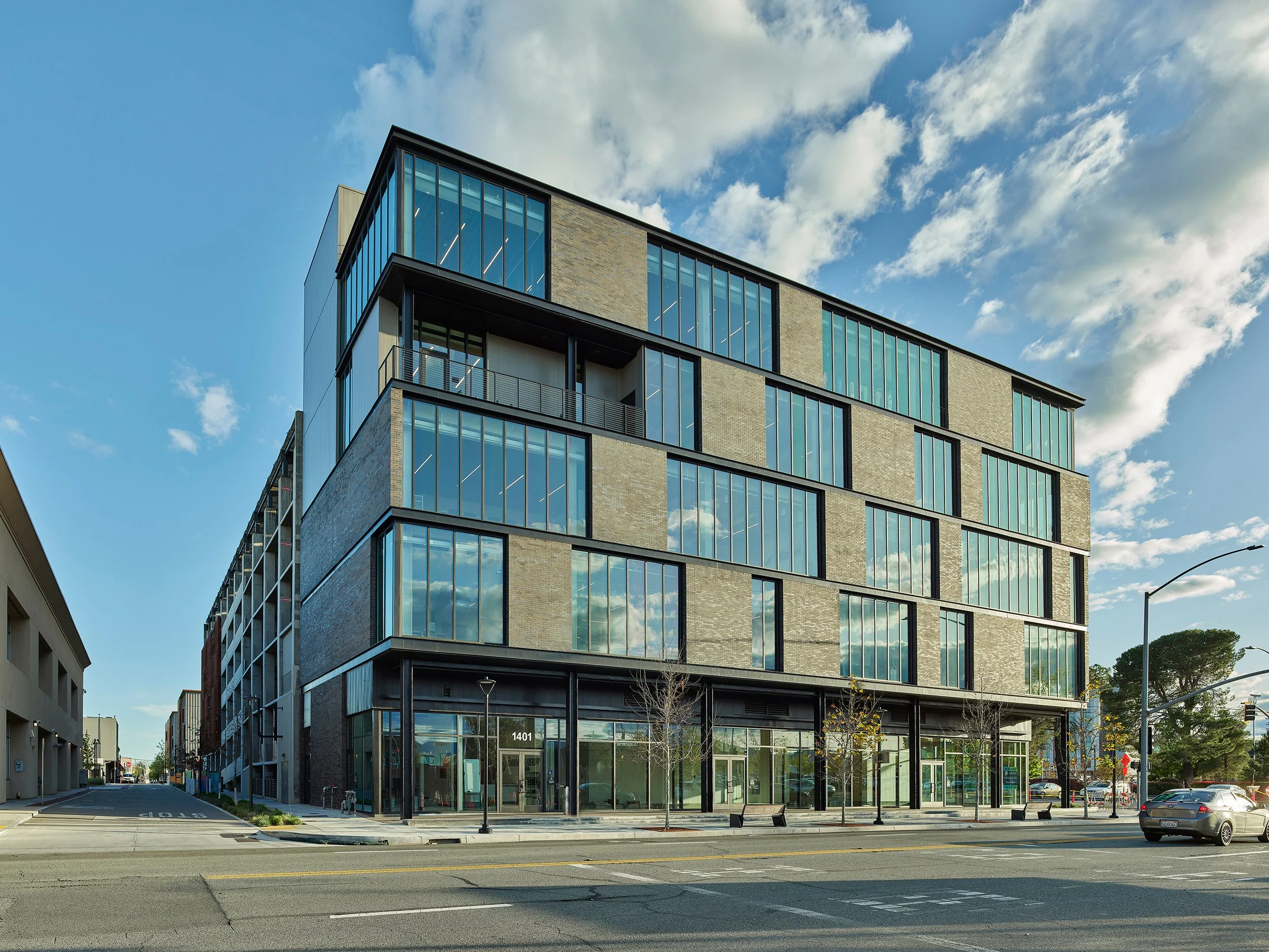 Shasta College in Redding, California. Part of Block 7 Development. A modern multi-story building with large glass windows reflecting the sky with clouds, some trees, and cars parked on the street.
