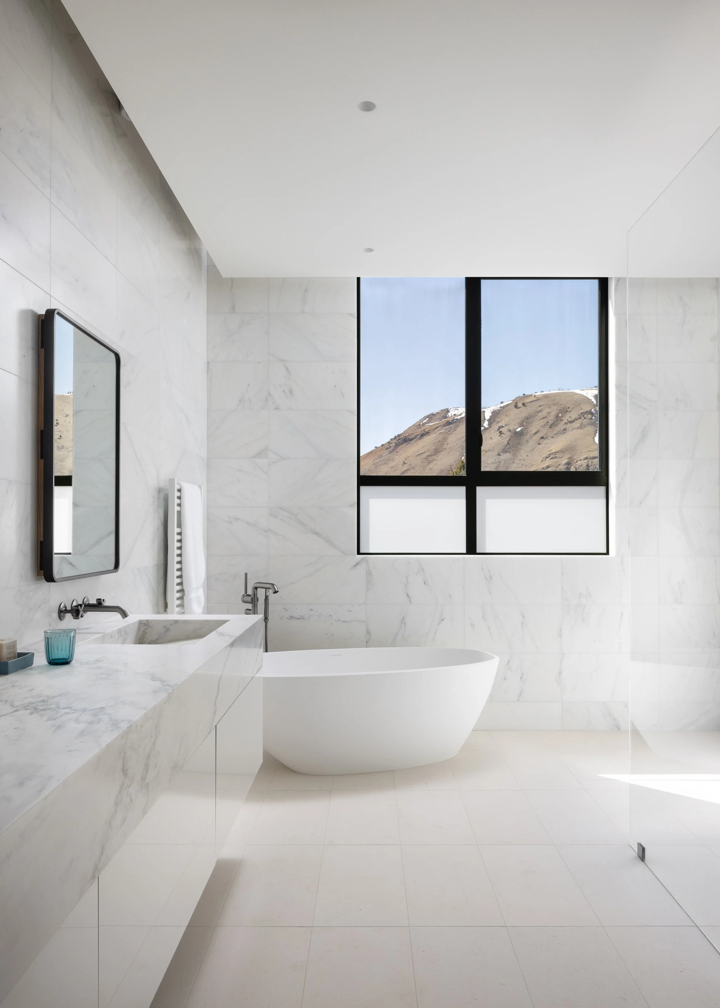 Split Timber House in Jackson, Wyoming. A modern bathroom with white marble walls and flooring, a freestanding white bathtub, a marble vanity with a built-in sink, and a large window showing mountain scenery outside.