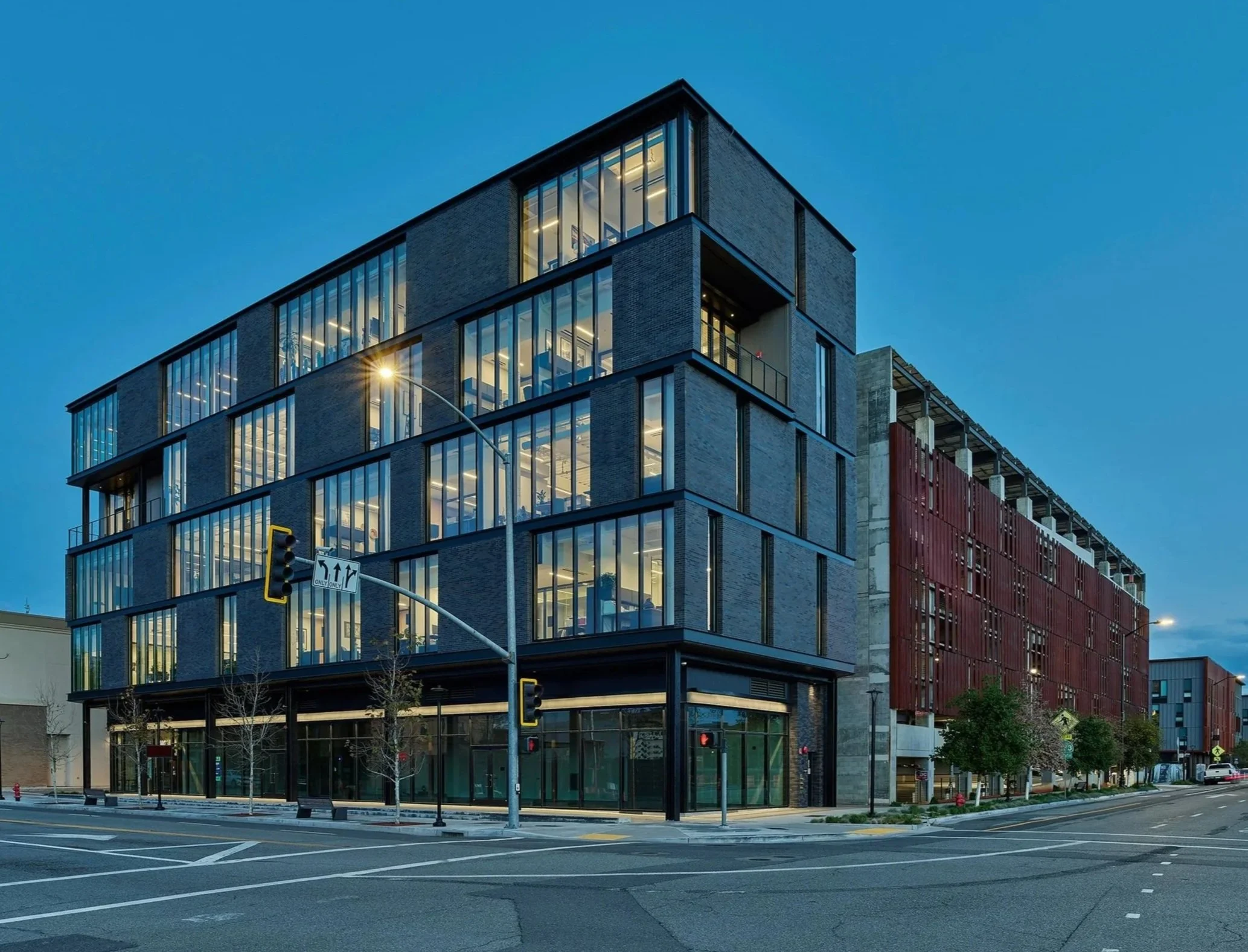 Shasta College in Redding, California. Part of Block 7 Development. A modern building with large glass windows and a brick exterior, lit from within, situated at a street corner with traffic lights and street signs, during early evening