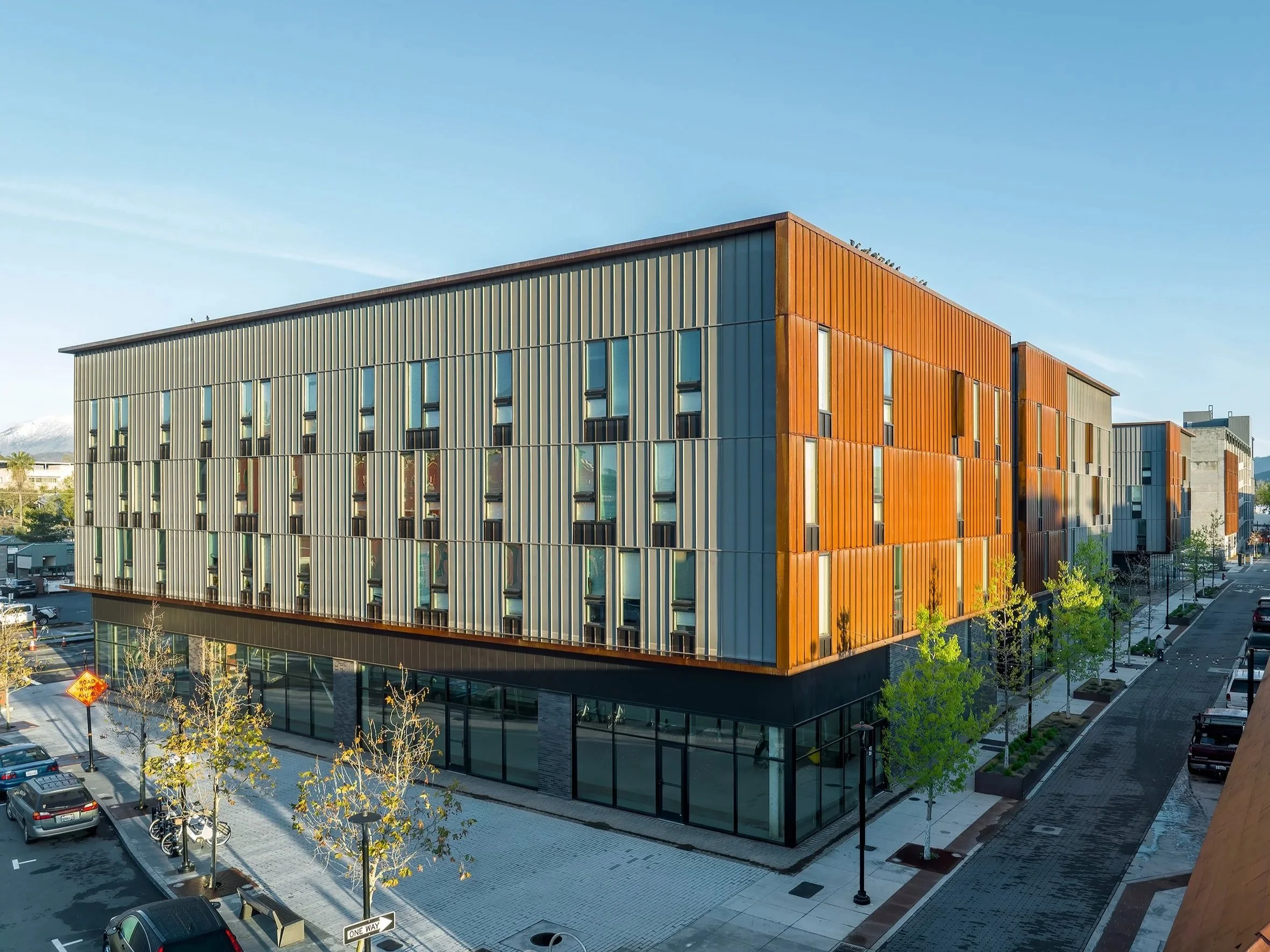 Block 7 Development in Redding, California. Modern multi-story building with vertical orange and beige siding, glass windows, and a parking lot with cars and young trees in front.