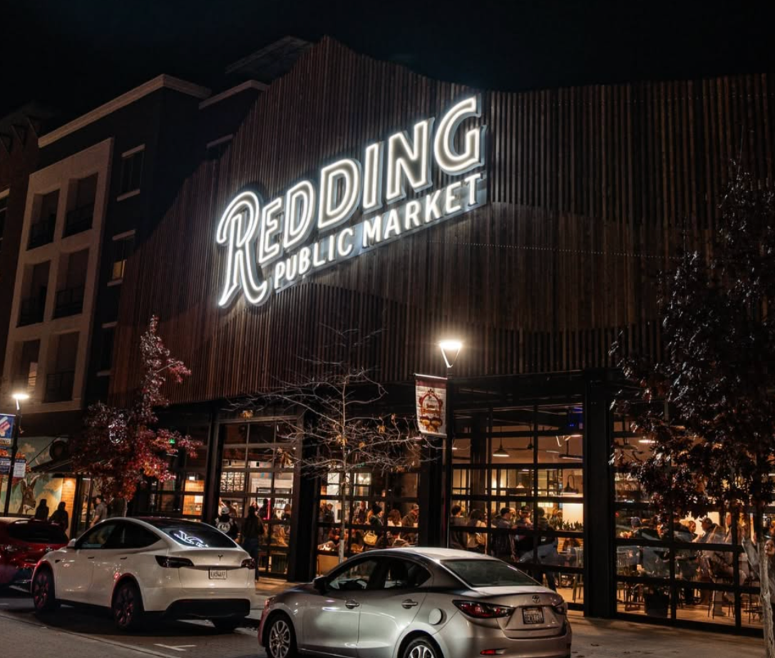 Night view of Redding Public Market with illuminated sign, cars parked outside, and people inside the market.